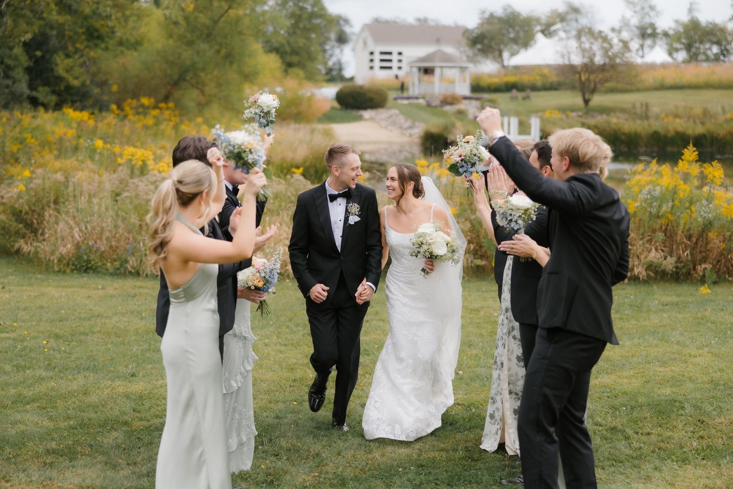 Wedding party walks through golden field as bridesmaids in sage gowns raise bouquets to celebrate newlywed couple.