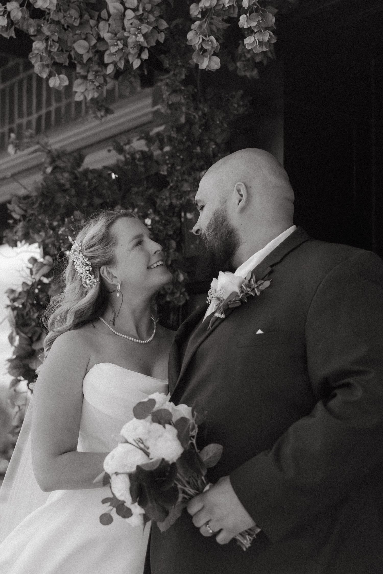 Black and white wedding photo of smiling bride holding bouquet gazing up at bearded groom in suit outdoors.