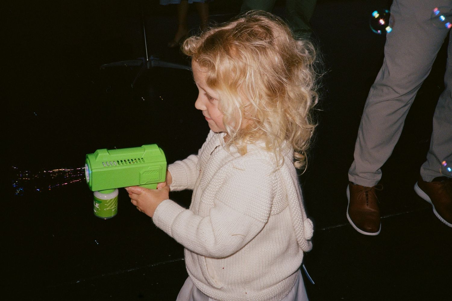 Adorable toddler girl with curly blonde hair playing with a green bubble gun at a wedding reception.