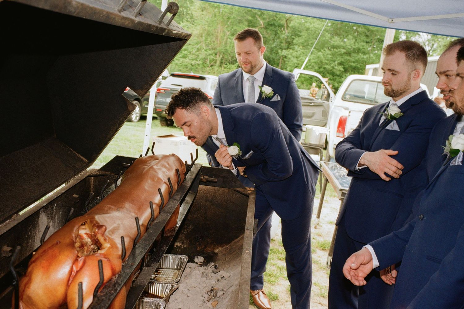 Groom in navy suit leaning over a whole roasted pig on a spit grill surrounded by groomsmen at wedding.