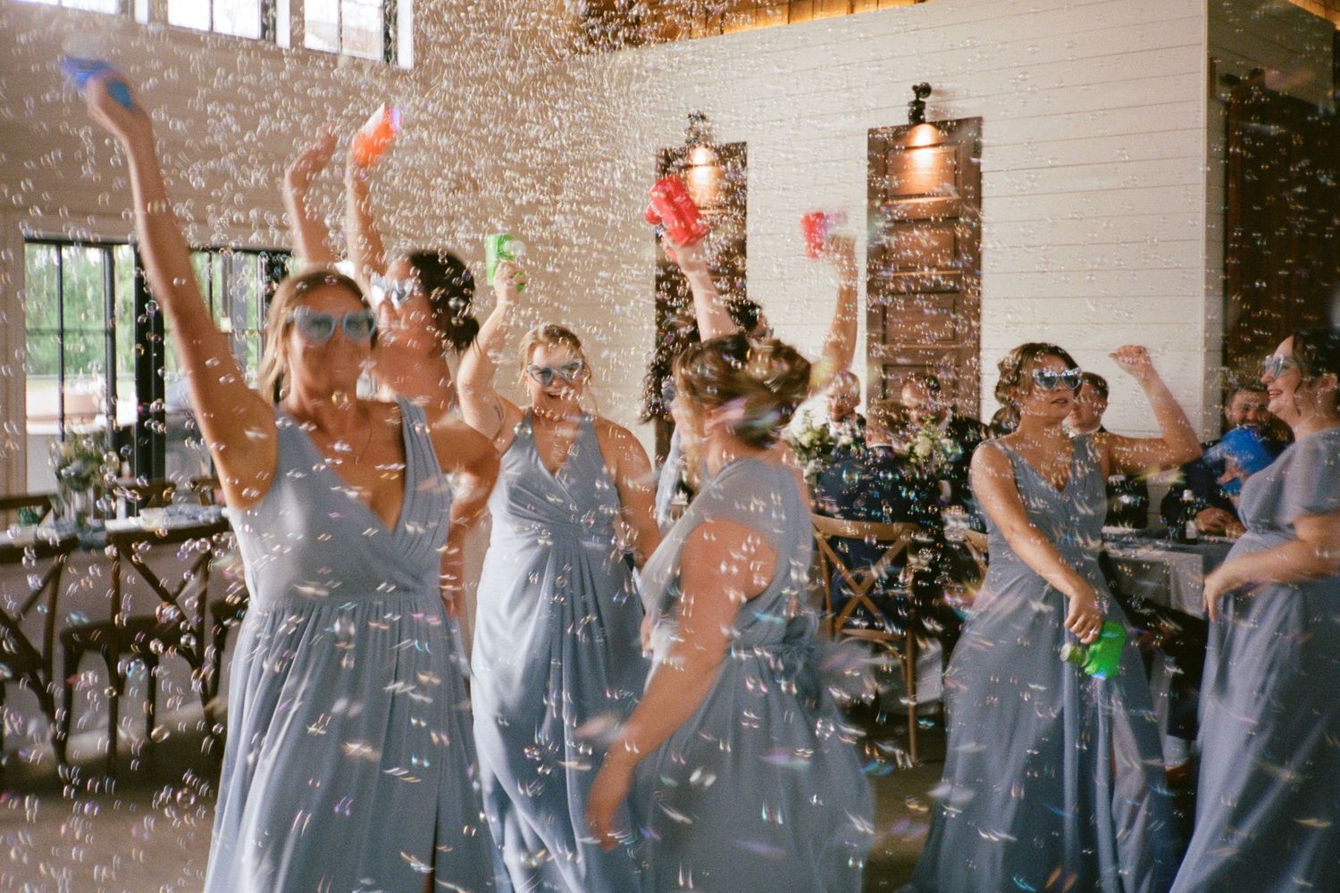 Bridesmaids in blue-gray dresses dancing with confetti and bubbles at a festive indoor wedding reception.