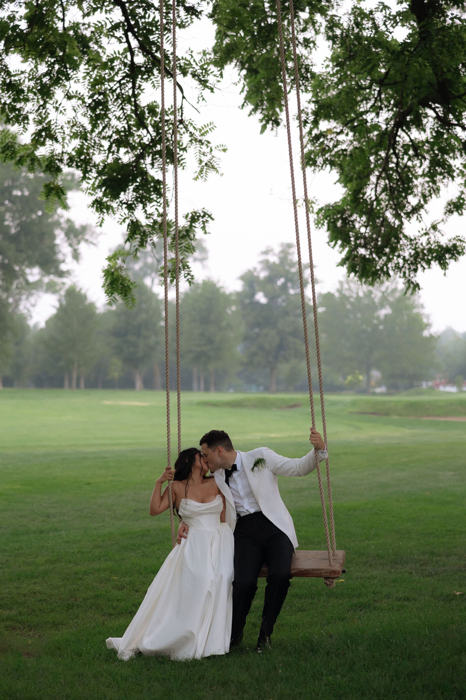 Bride and groom sharing a romantic kiss on a wooden swing hanging from a tree in a lush green outdoor setting.