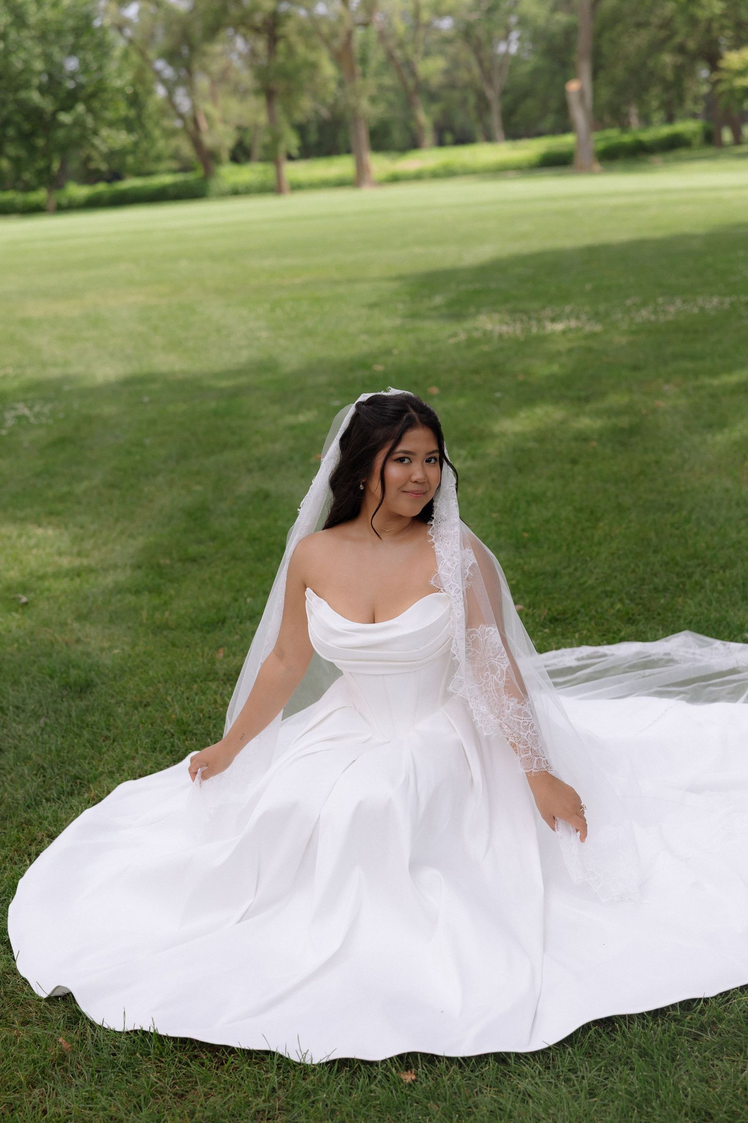 Beautiful bride in a strapless white gown with lace veil sitting gracefully on green grass.