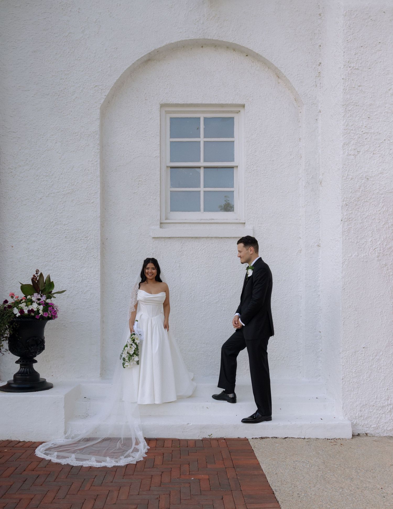 Bride and groom sharing a first look moment beside a white arched building with brick pathway.