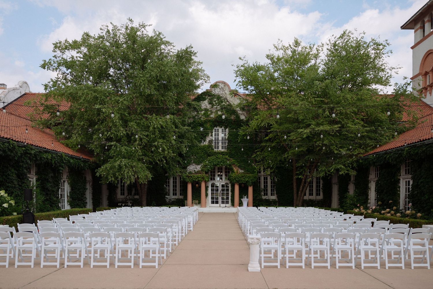 Outdoor wedding ceremony setup with white chairs arranged in rows on a patio, framed by lush trees and an elegant ivy-covered venue.