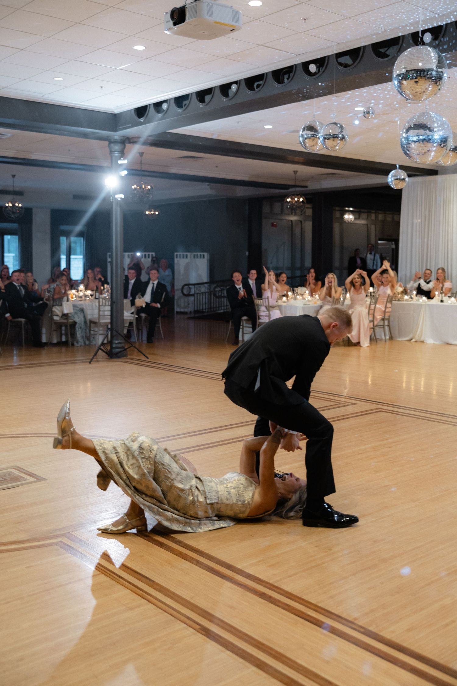 Dramatic wedding dance dip with groom bending over bride lying on ballroom floor before guests.