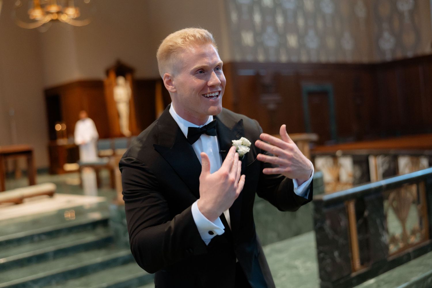 Smiling blond groom in black tuxedo gestures expressively inside an elegant church before the ceremony.