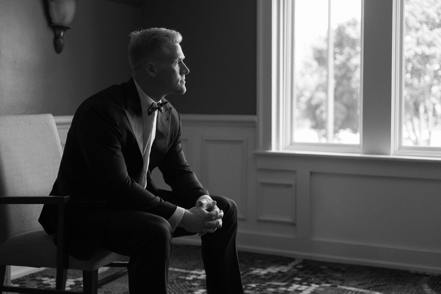 Black and white photo of groom in tuxedo sitting pensively by window.