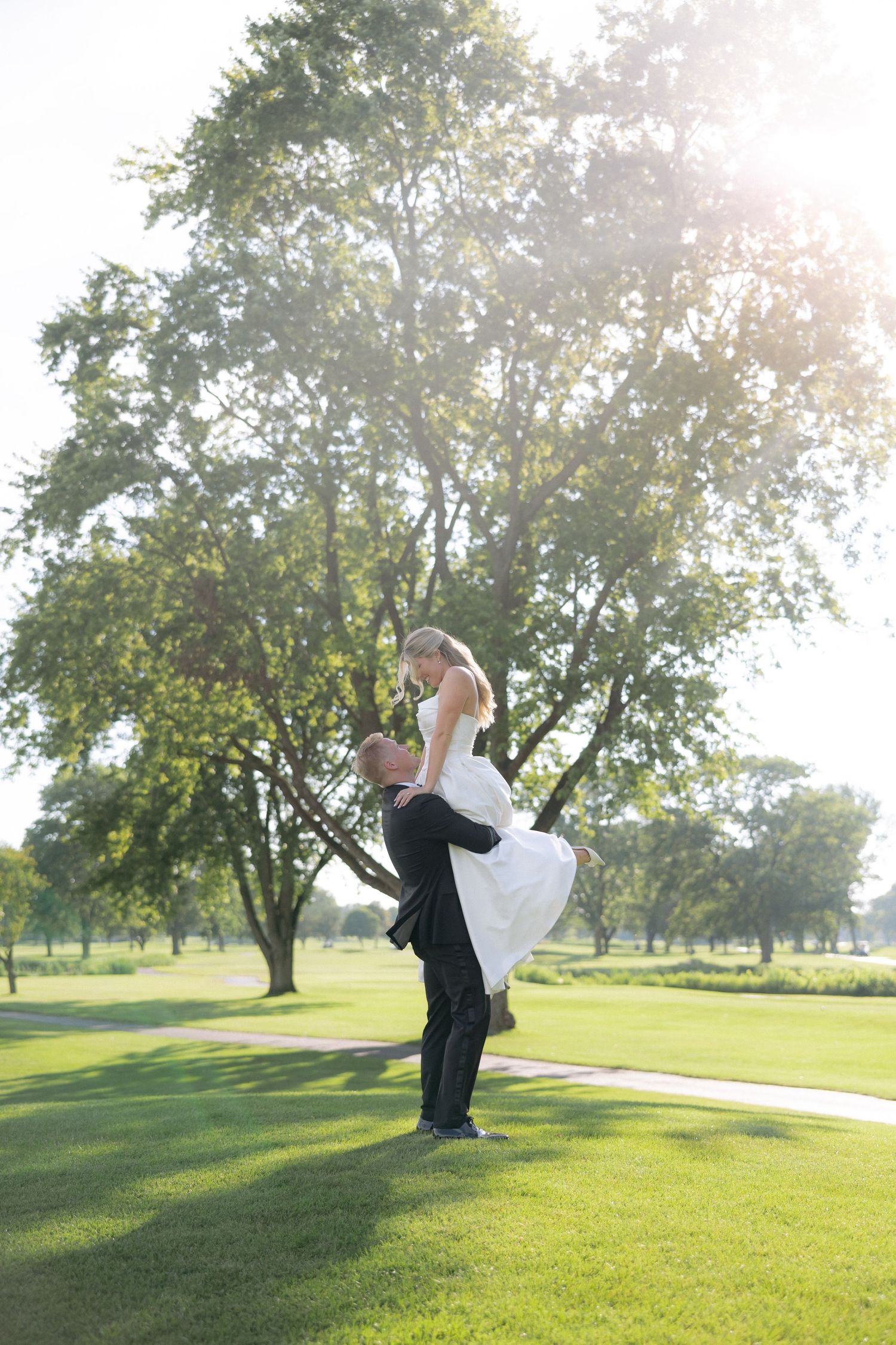 Groom lifts bride joyfully in sunlit park with large green trees in background.