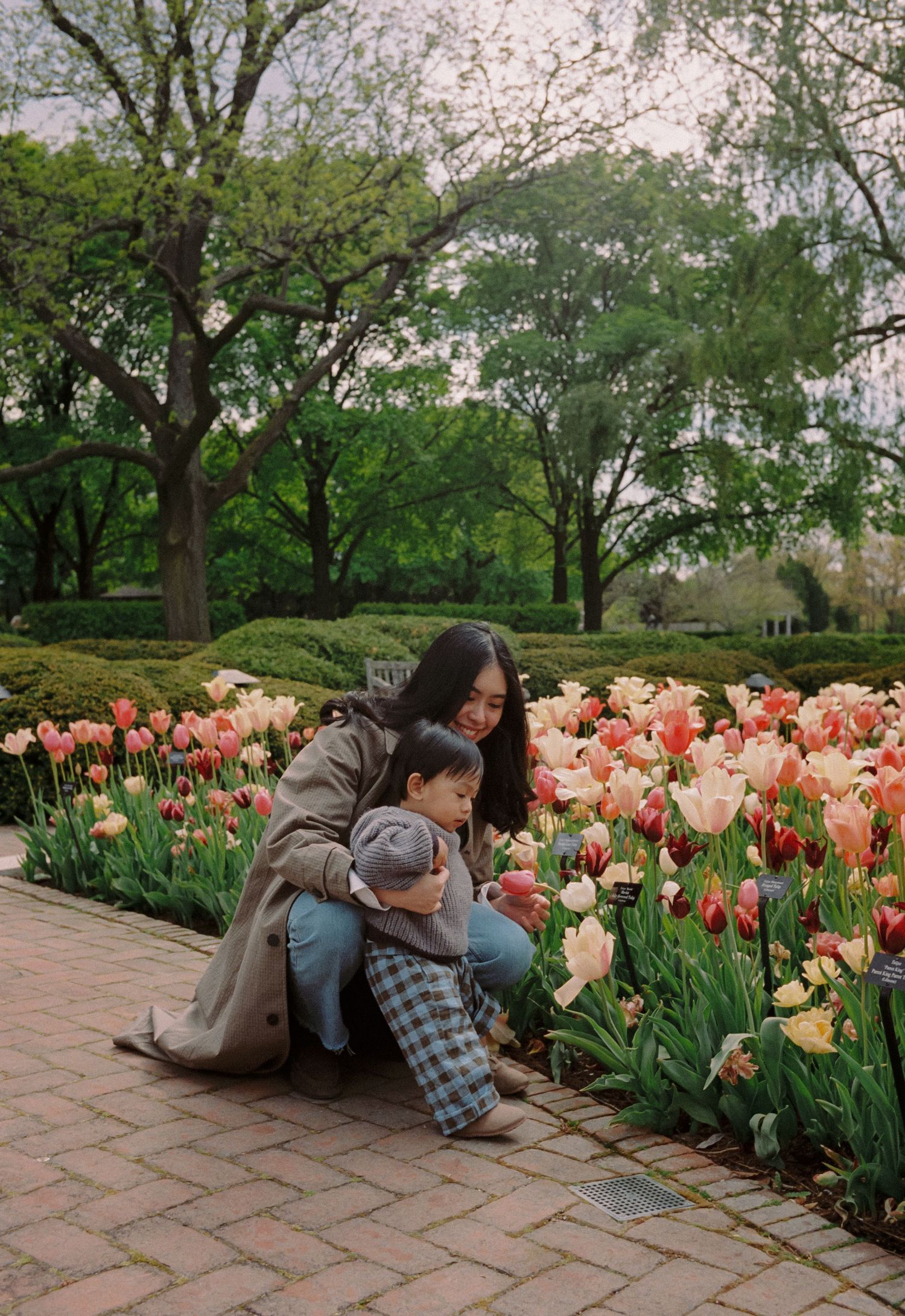 A woman and toddler crouch beside colorful tulips in a lush garden park during spring bloom.