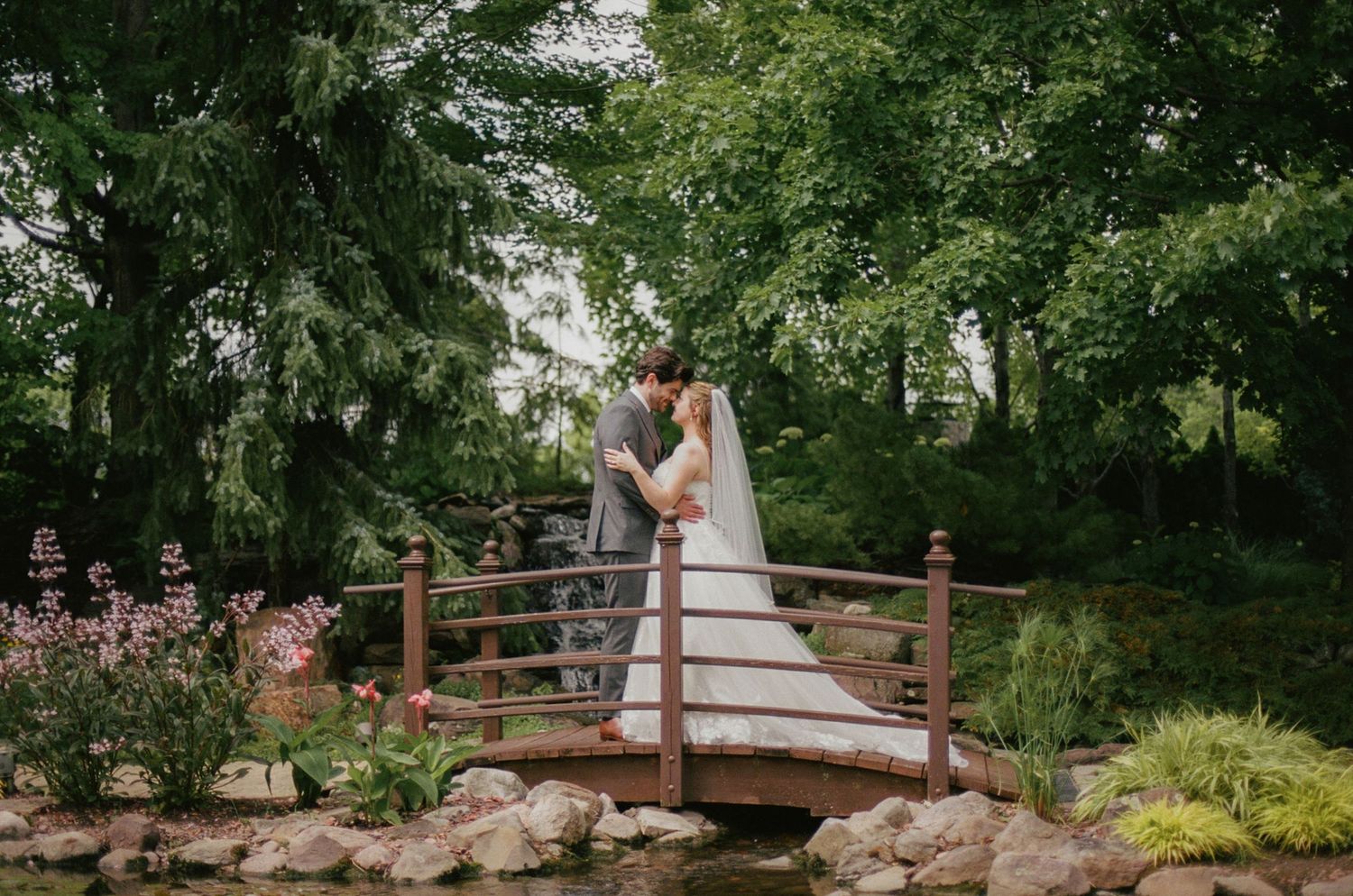 Bride and groom kissing on a wooden bridge surrounded by lush green trees in a romantic garden wedding setting.