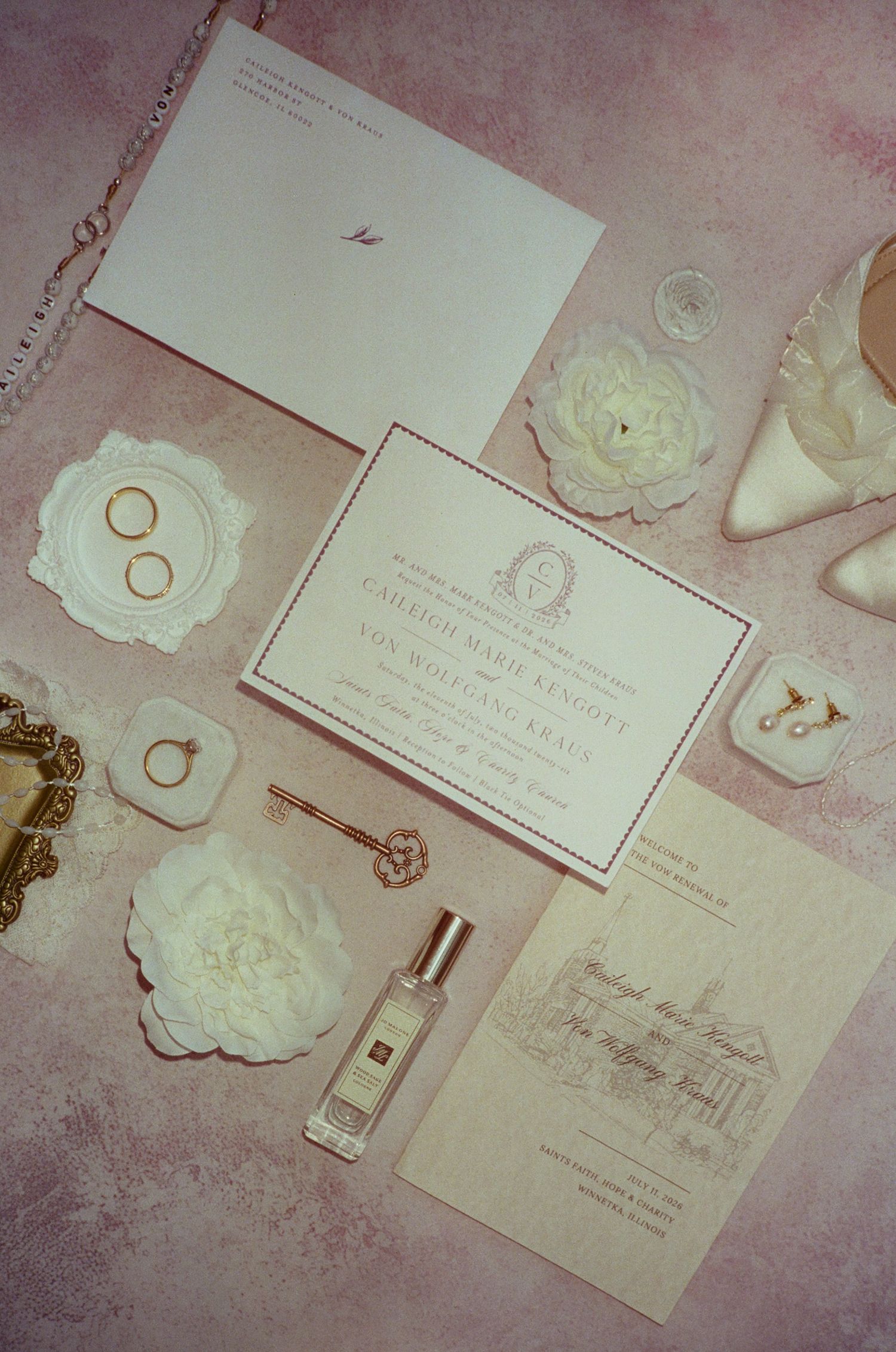 Elegant wedding flat lay with floral invitations, gold rings, Jo Malone perfume, white peonies, and bridal heels on pink background.