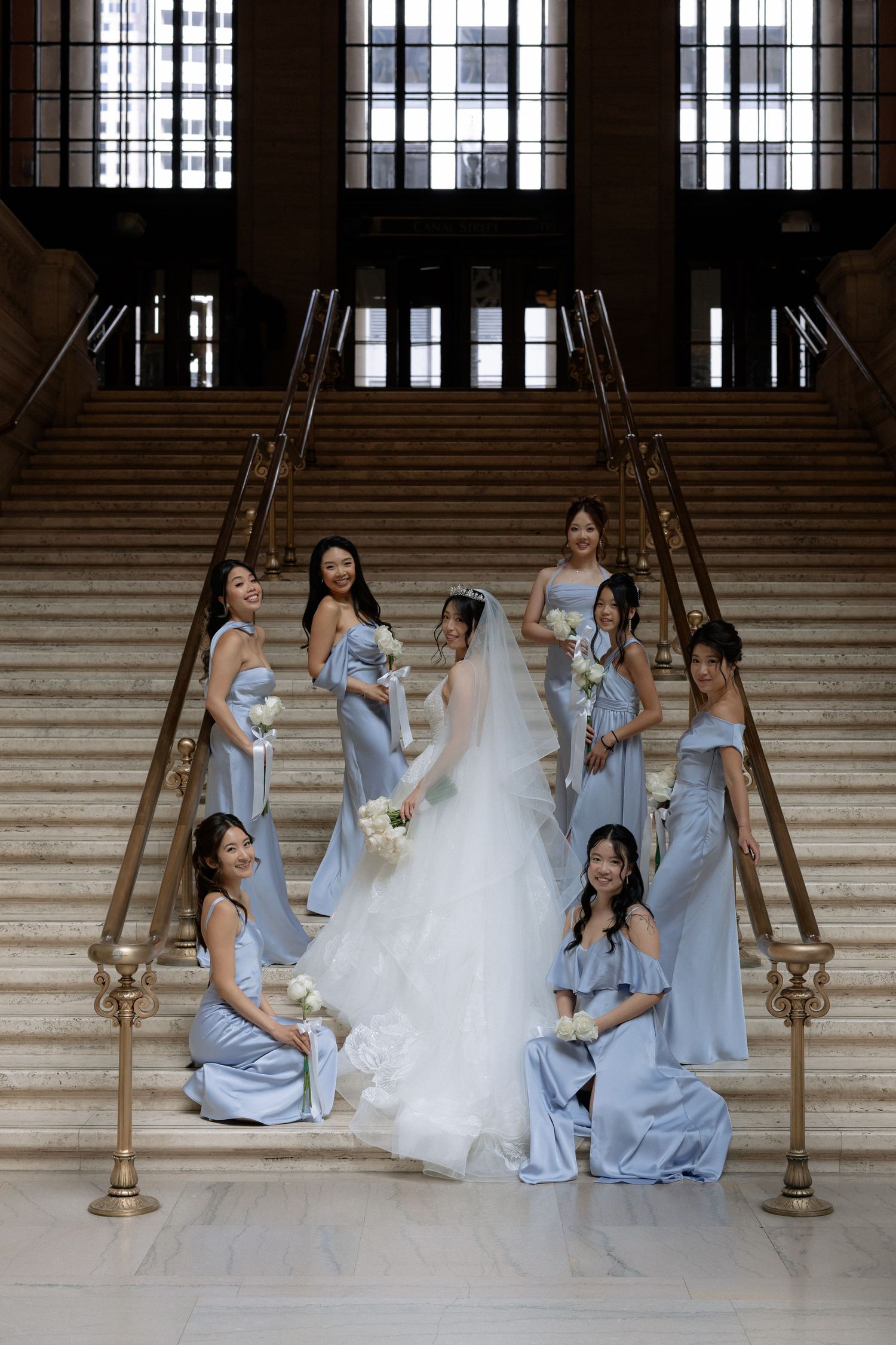 Bride in white gown surrounded by bridesmaids in blue dresses posing on grand staircase.