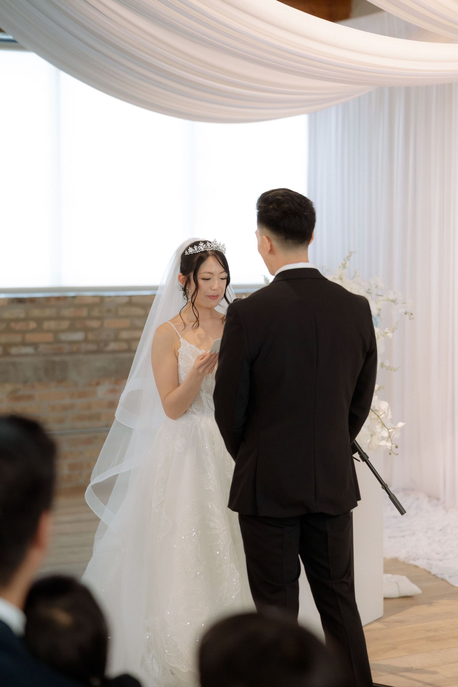 Bride and groom exchange vows at elegant wedding ceremony with white draping and floral decor.