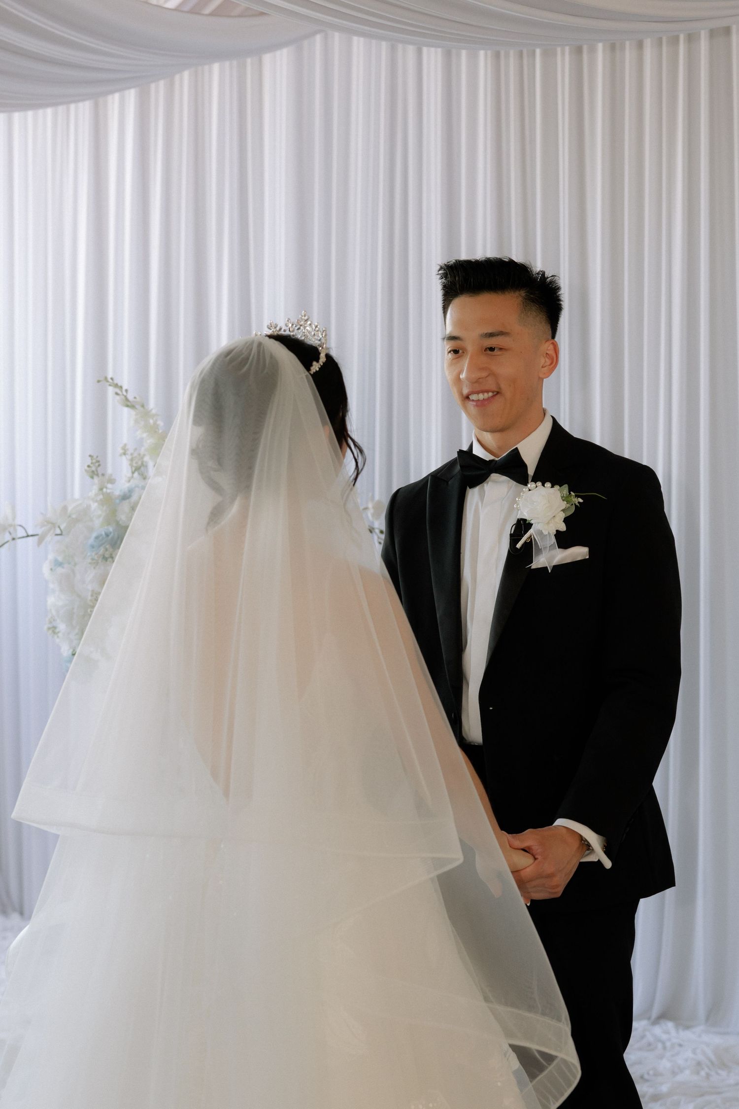 Smiling groom in black tuxedo holds bride's hands at altar during elegant white wedding ceremony.
