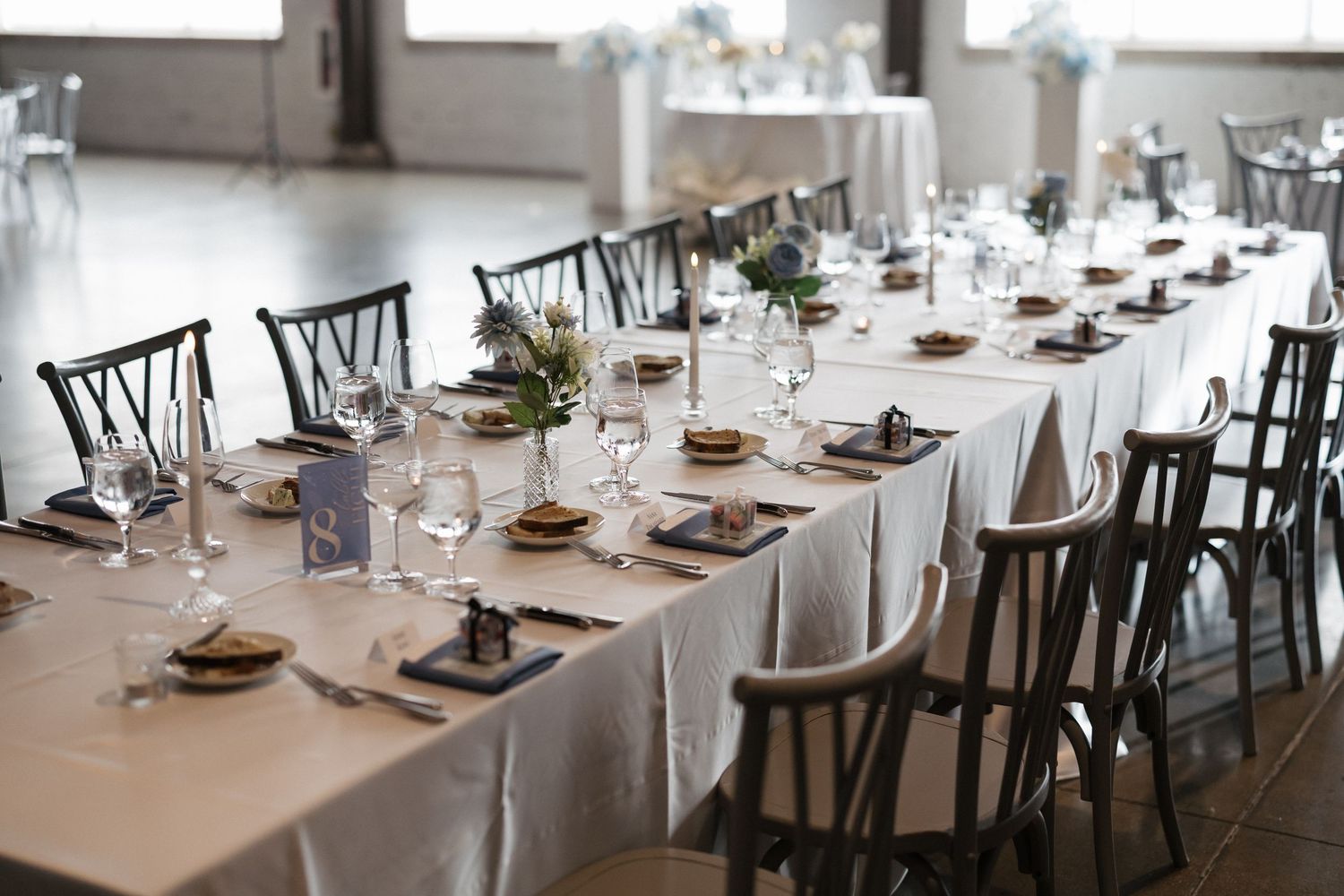 Elegant banquet table set with white linens, dark chairs, glassware, and floral centerpieces in a bright reception hall.