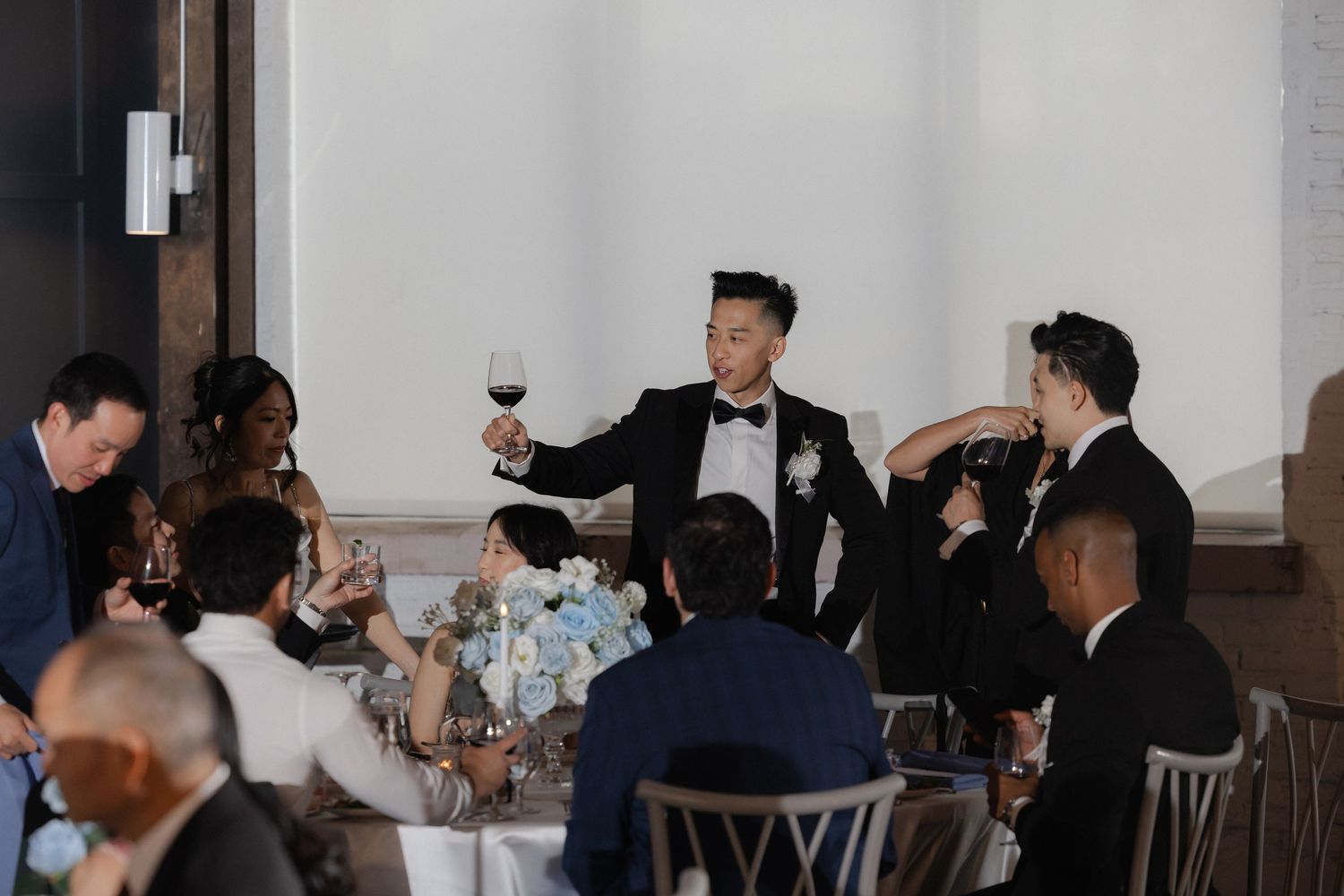 Groom in tuxedo raises wine glass for toast surrounded by wedding guests at elegant reception dinner.