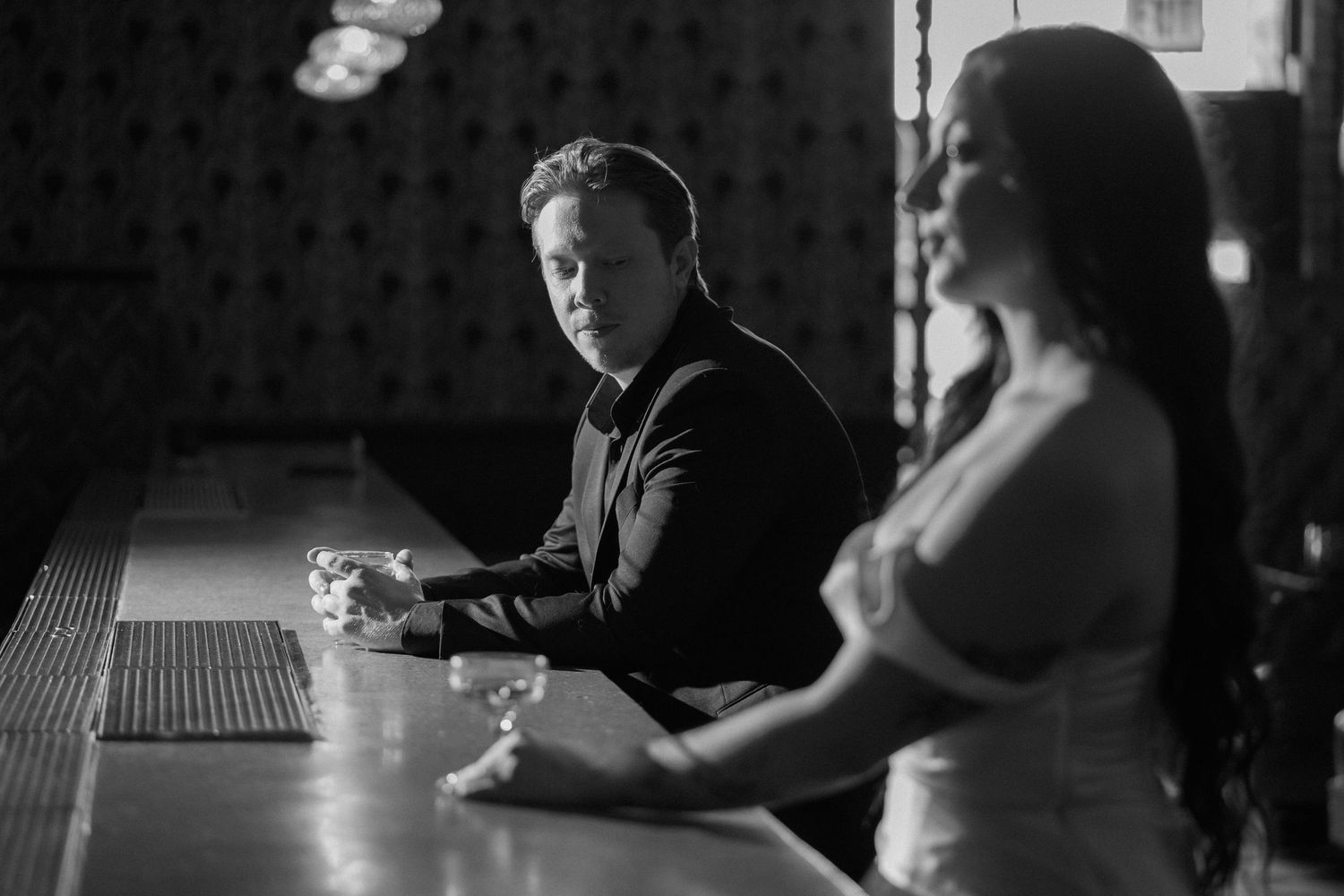 Black and white photo of a man in a black shirt sitting at a bar looking at a woman in white dress.