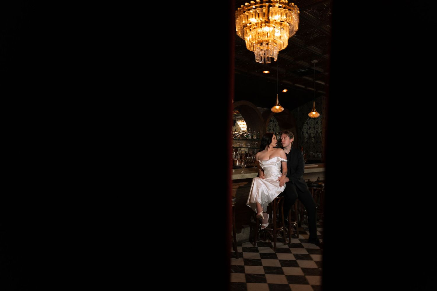 Bride and groom share a romantic moment under a chandelier in a dimly lit elegant bar with checkered floors.