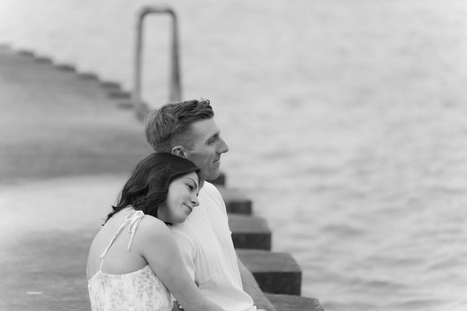 Black and white photo of couple sitting together on steps by calm waterfront.