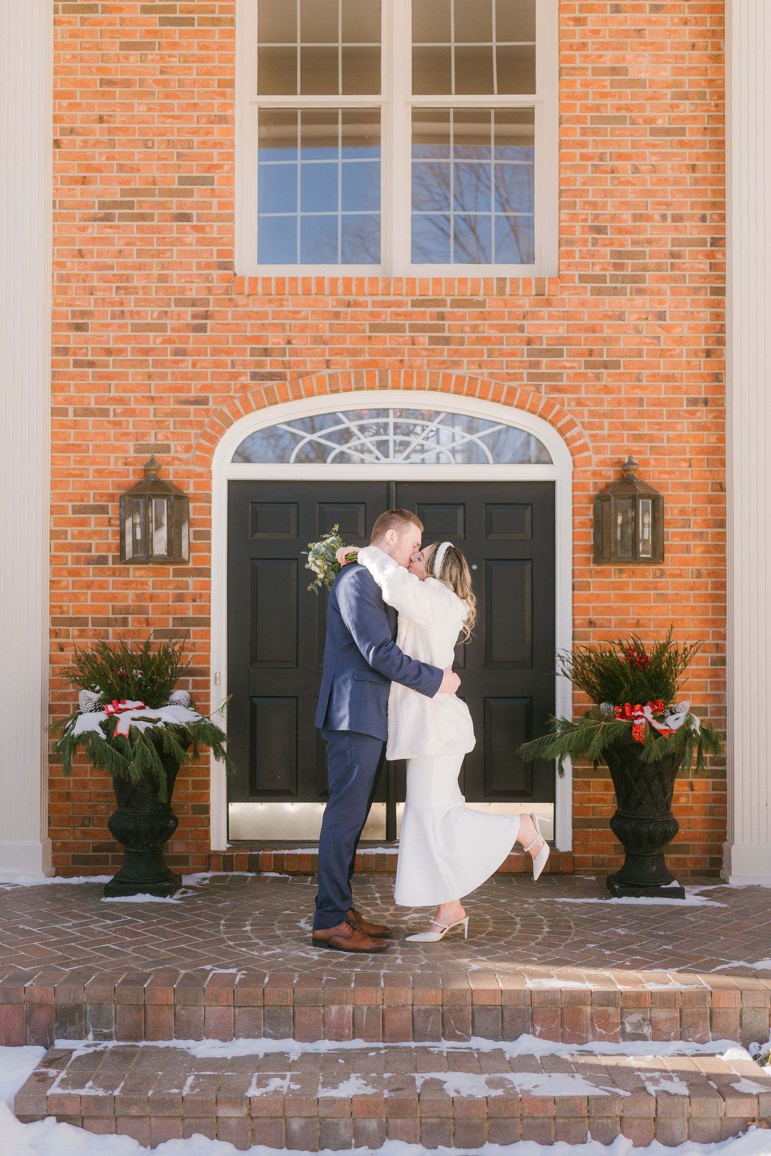 Newlyweds share a romantic kiss in front of brick mansion entrance decorated for winter holidays.