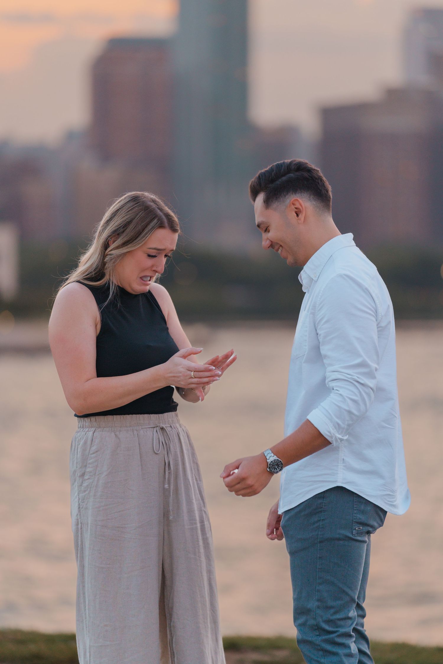 Man presenting an engagement ring to an emotional woman by a waterfront at sunset.