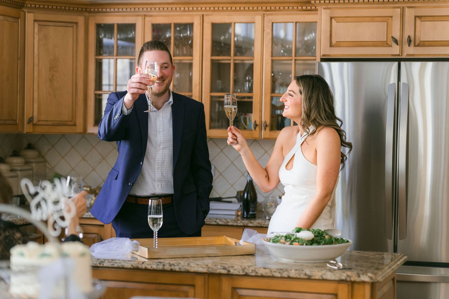 Bride and groom laughing while toasting champagne glasses together in a warmly lit kitchen.