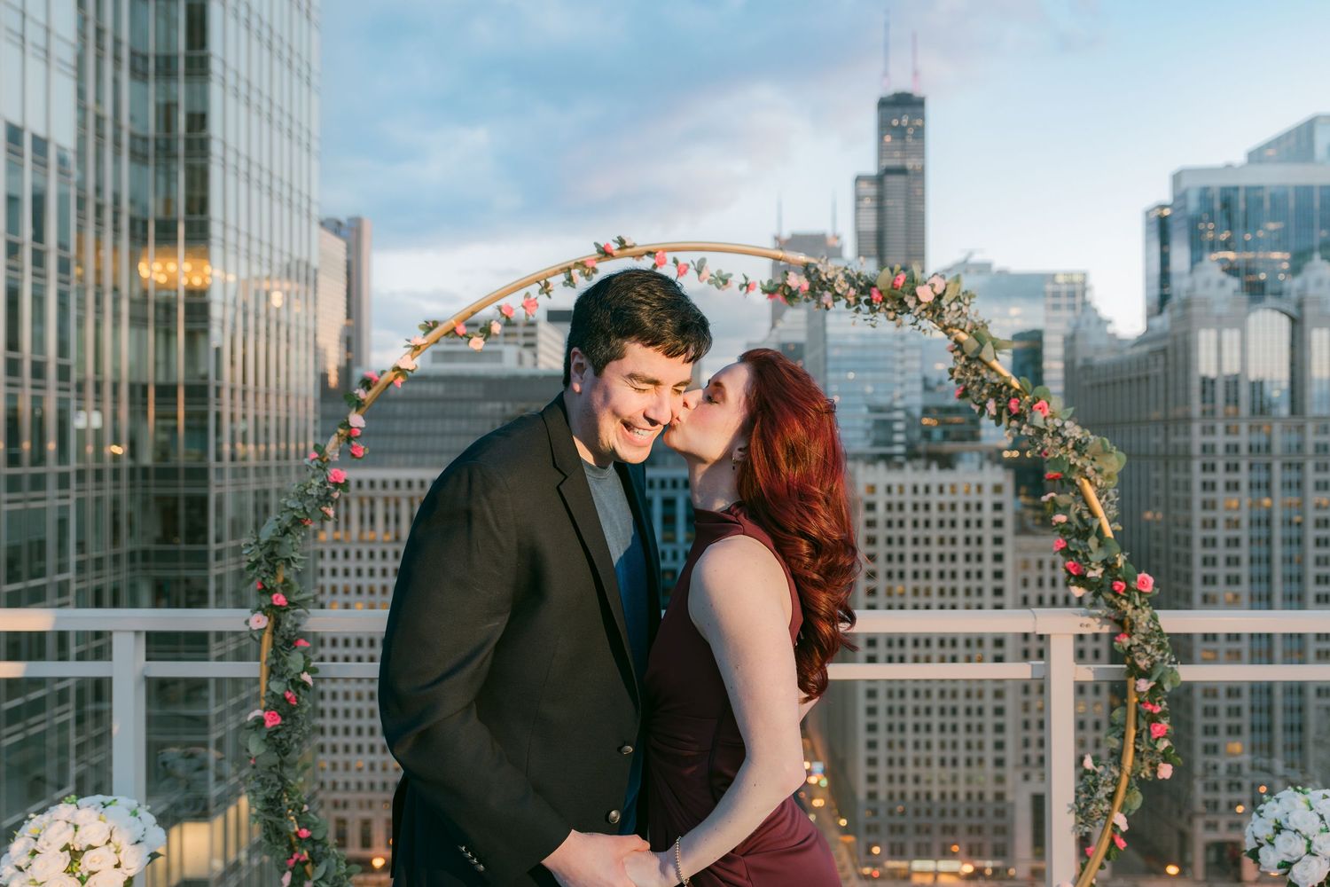 Happy couple sharing a kiss under a floral hoop arch with Chicago skyline at golden hour.