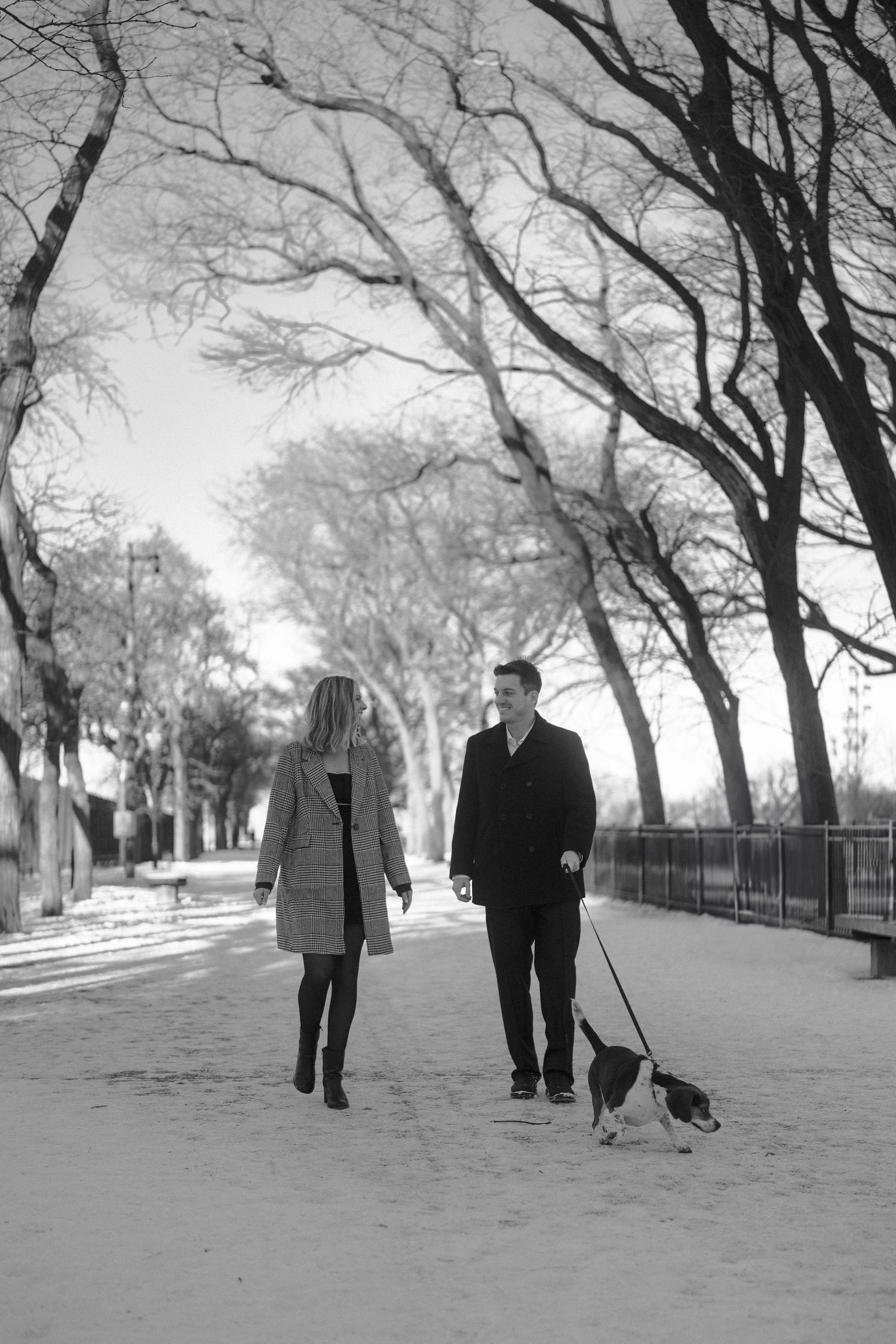 Couple walking a beagle dog along a snowy tree-lined path in black and white photo.