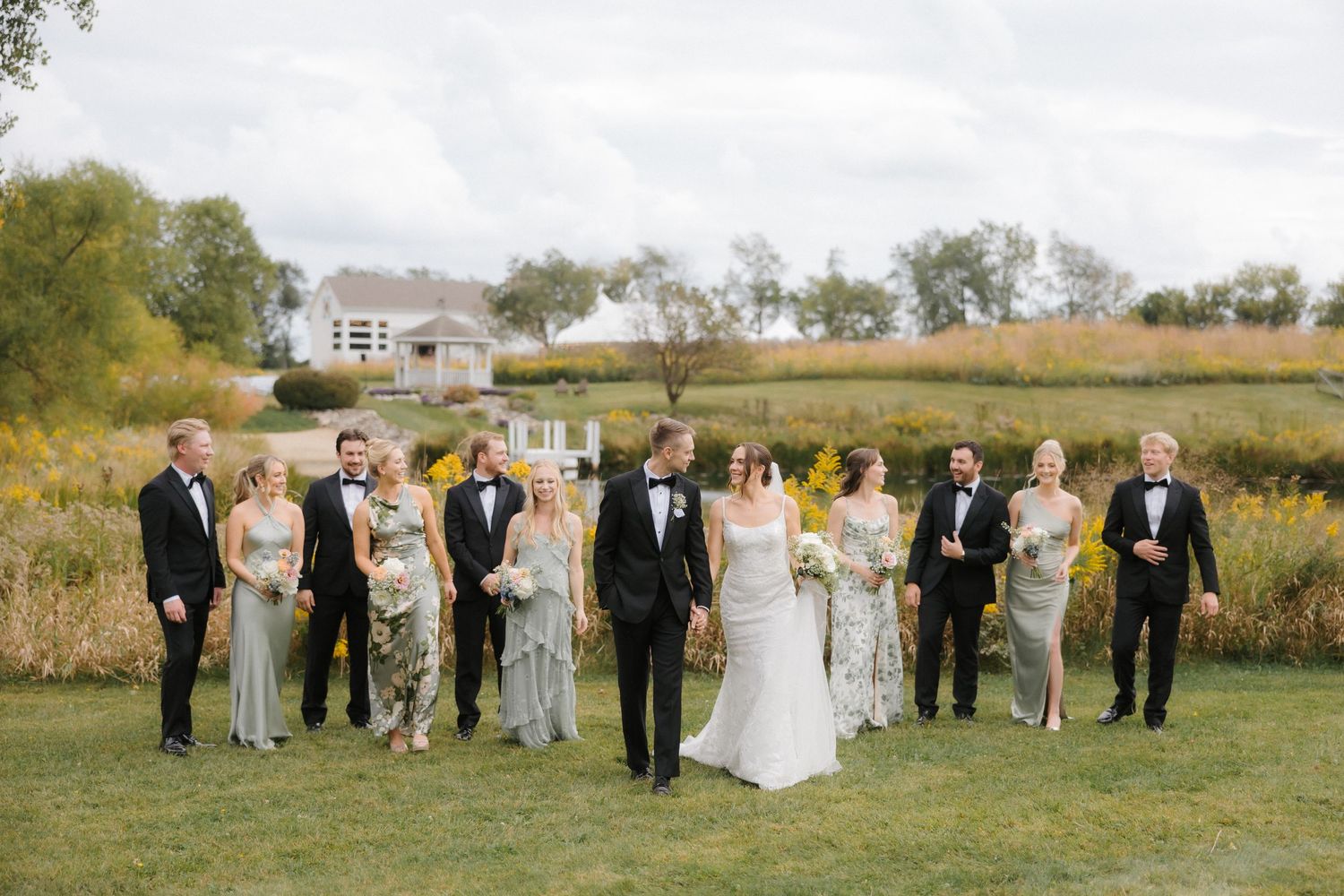 Wedding party strolls across green lawn in black tuxedos and sage dresses with a white farmhouse venue backdrop.