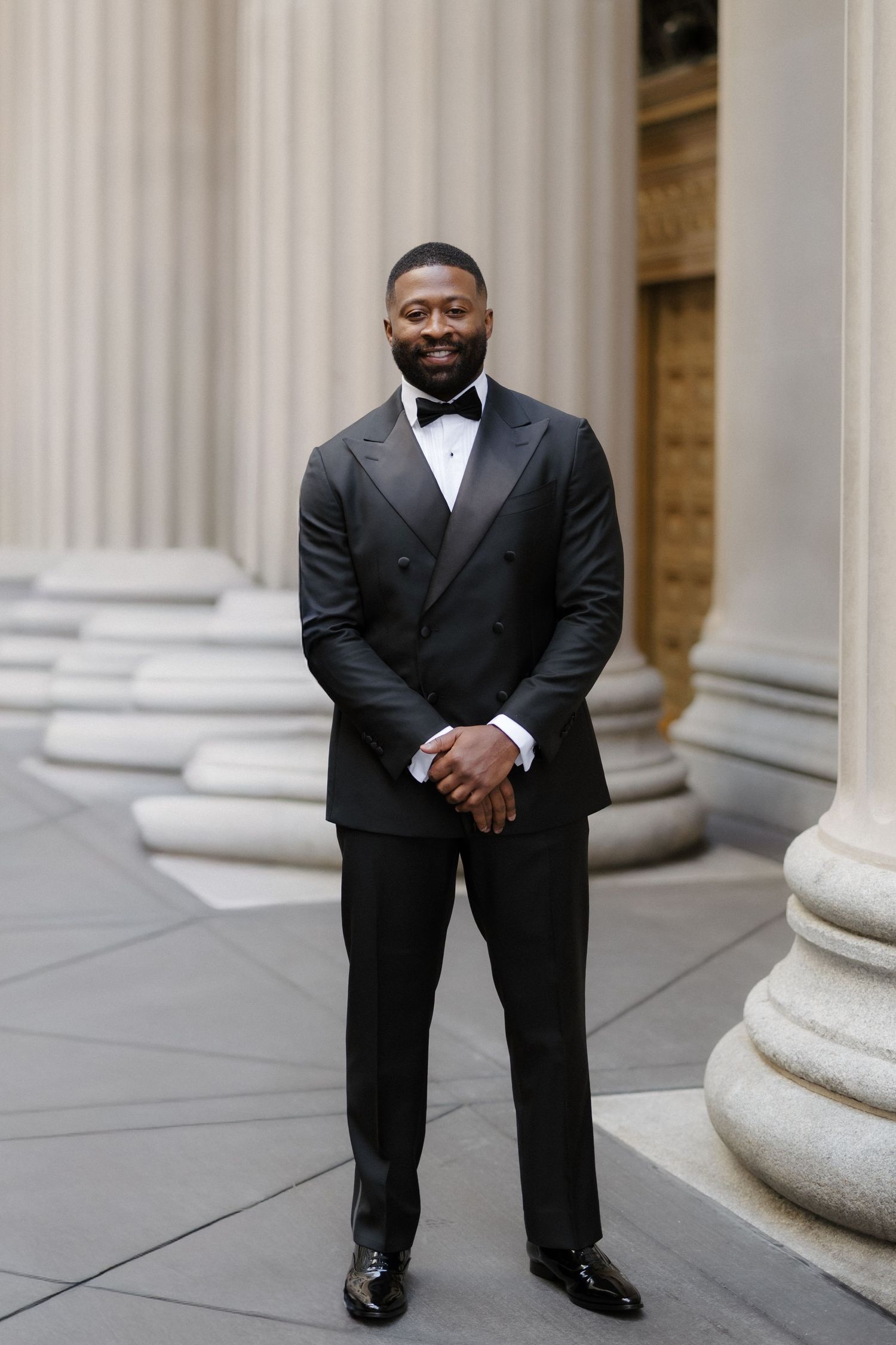 A well-dressed Black man in a black tuxedo with bow tie stands smiling between grand stone columns.