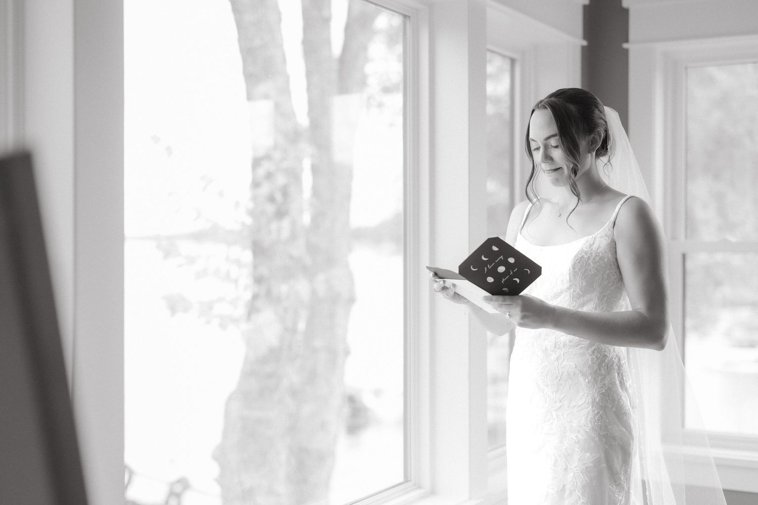 Black and white photo of bride in lace wedding dress smiling while reading a card by window.