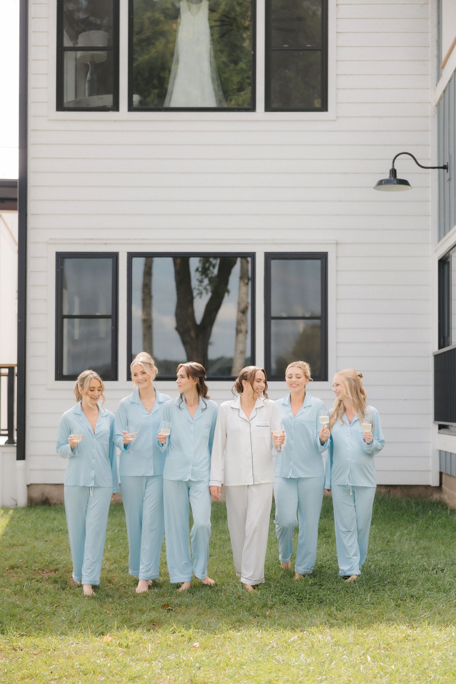 Bride and bridesmaids in matching light blue and white pajamas posing outside a white modern farmhouse.