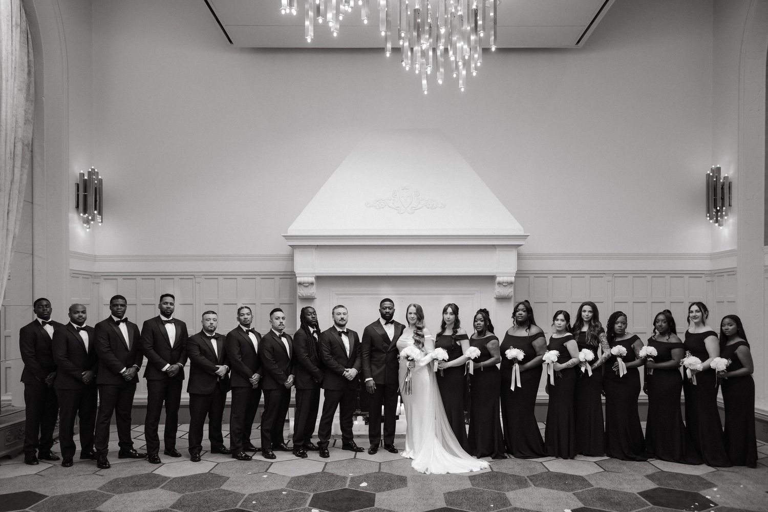 Black and white wedding party photo with groomsmen in tuxedos and bridesmaids in black gowns beside bride and groom in elegant venue.