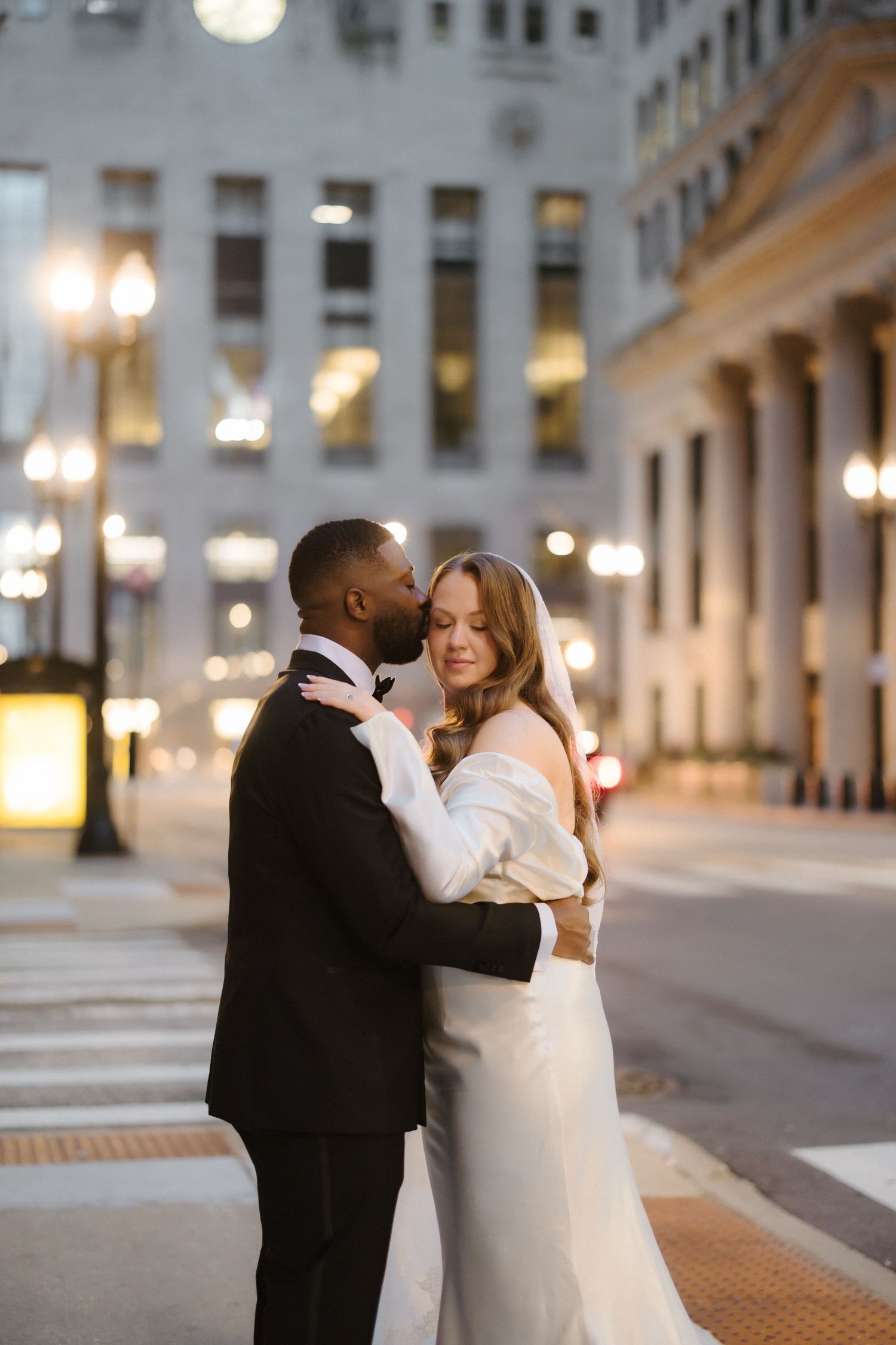 Interracial couple in wedding attire embrace on a city street at night, groom in black tuxedo kissing bride in white gown.