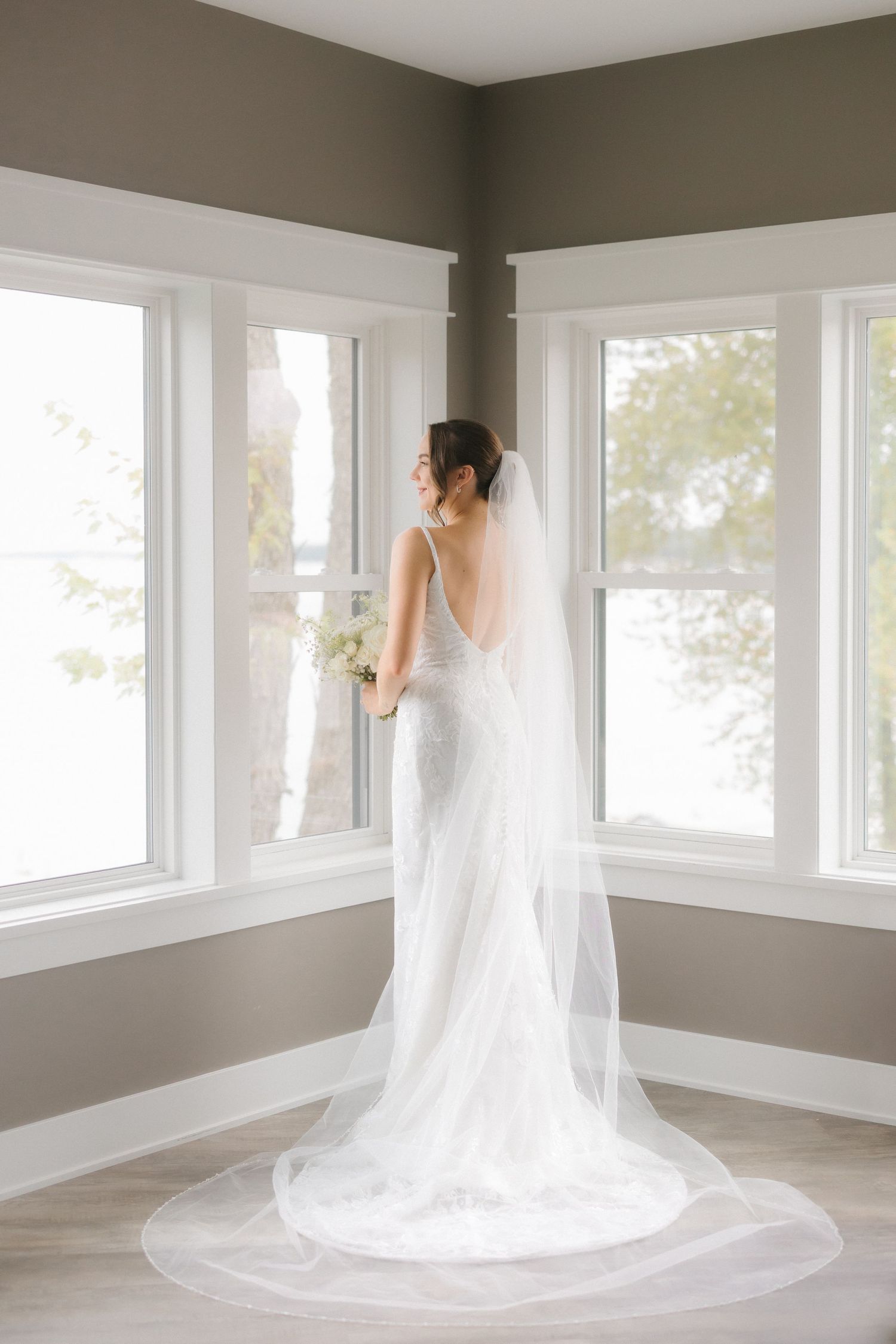 Bride in elegant lace gown with long cathedral veil holding white bouquet, viewed from behind near bright windows.