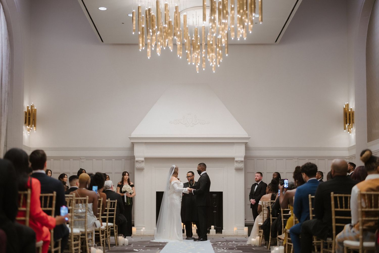 Elegant wedding ceremony in a ballroom with gold chandelier, couple at altar by white fireplace, guests seated in gold chairs.