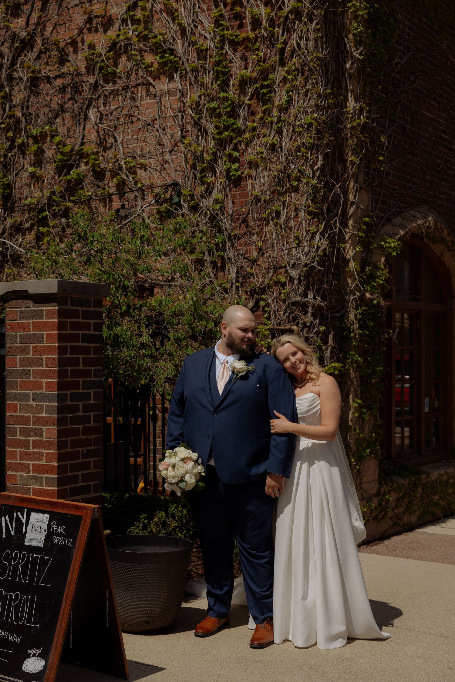 Bride and groom pose together outside ivy-covered brick wedding venue, bride holding white bouquet in elegant white gown.