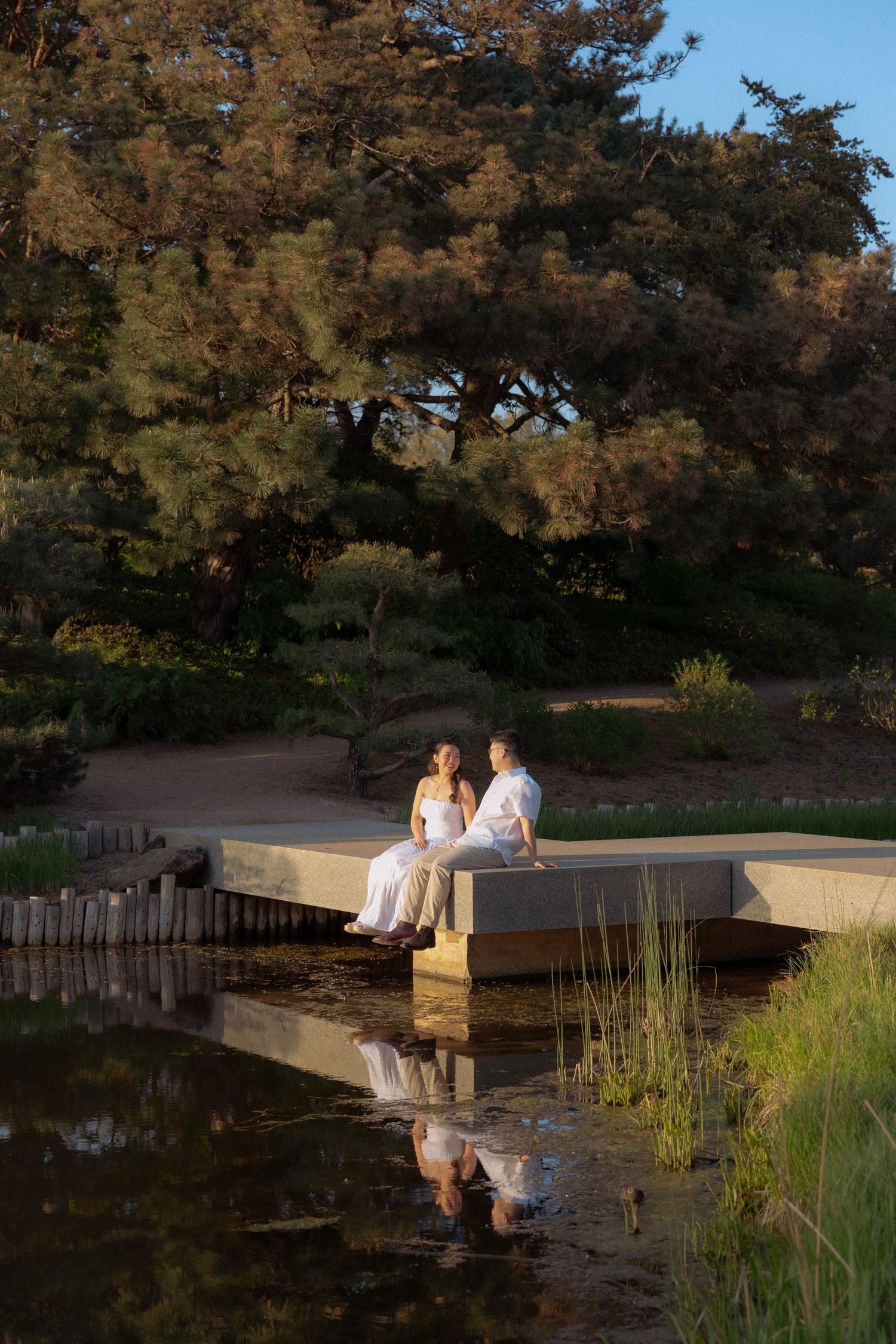 Couple in white sitting on a concrete bridge over a pond, reflected in water, surrounded by lush trees at golden hour.