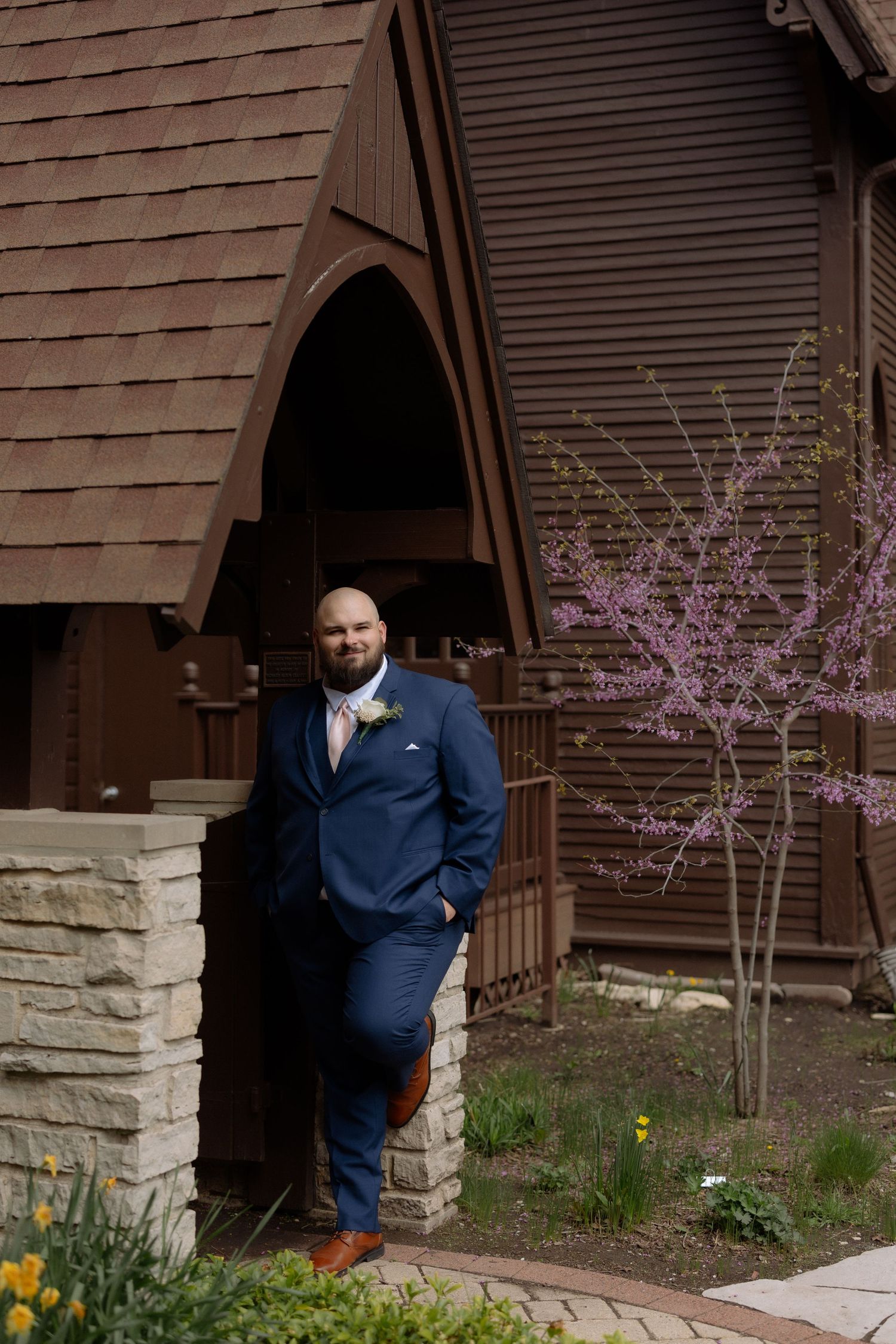 Groom in navy blue suit leaning against stone pillar outside rustic wooden chapel with spring blooming trees.