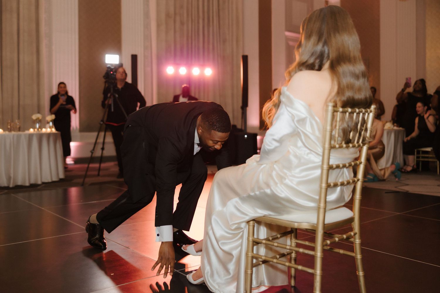Groom removing bride's garter at elegant wedding reception in grand ballroom with gold chiavari chairs.