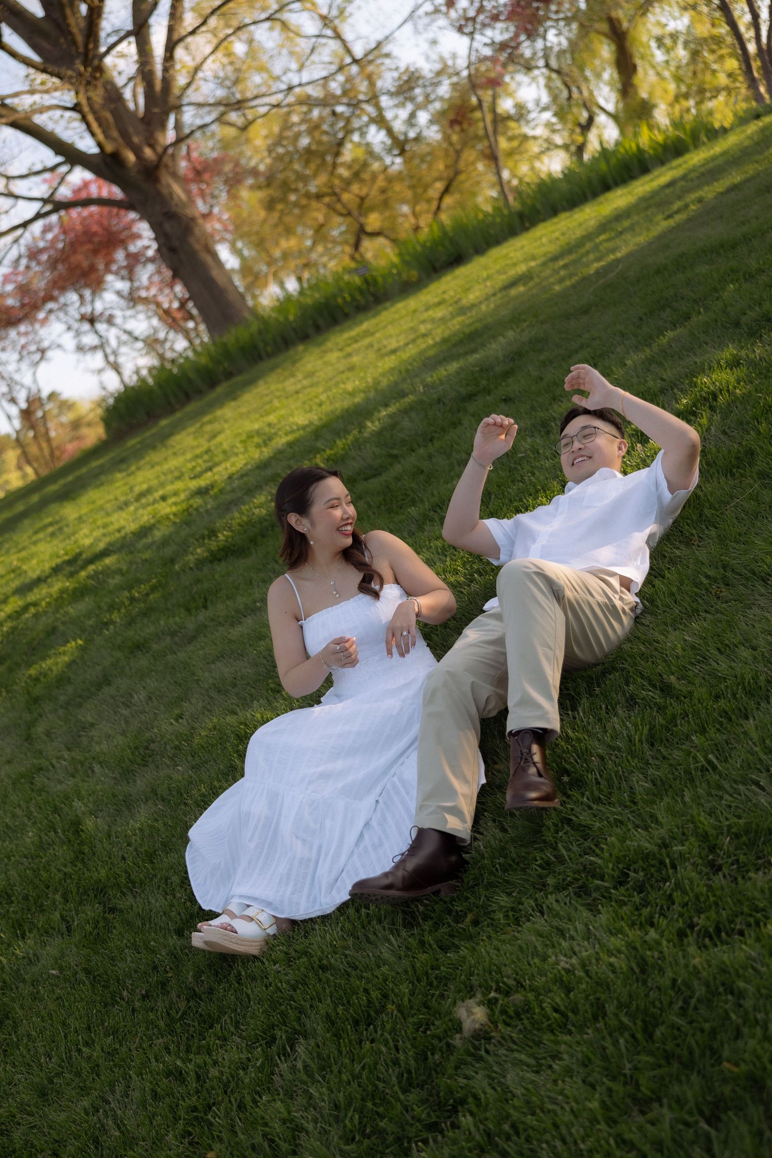 Happy Asian couple sitting on green grass hill in a park, laughing and enjoying a sunny day outdoors.
