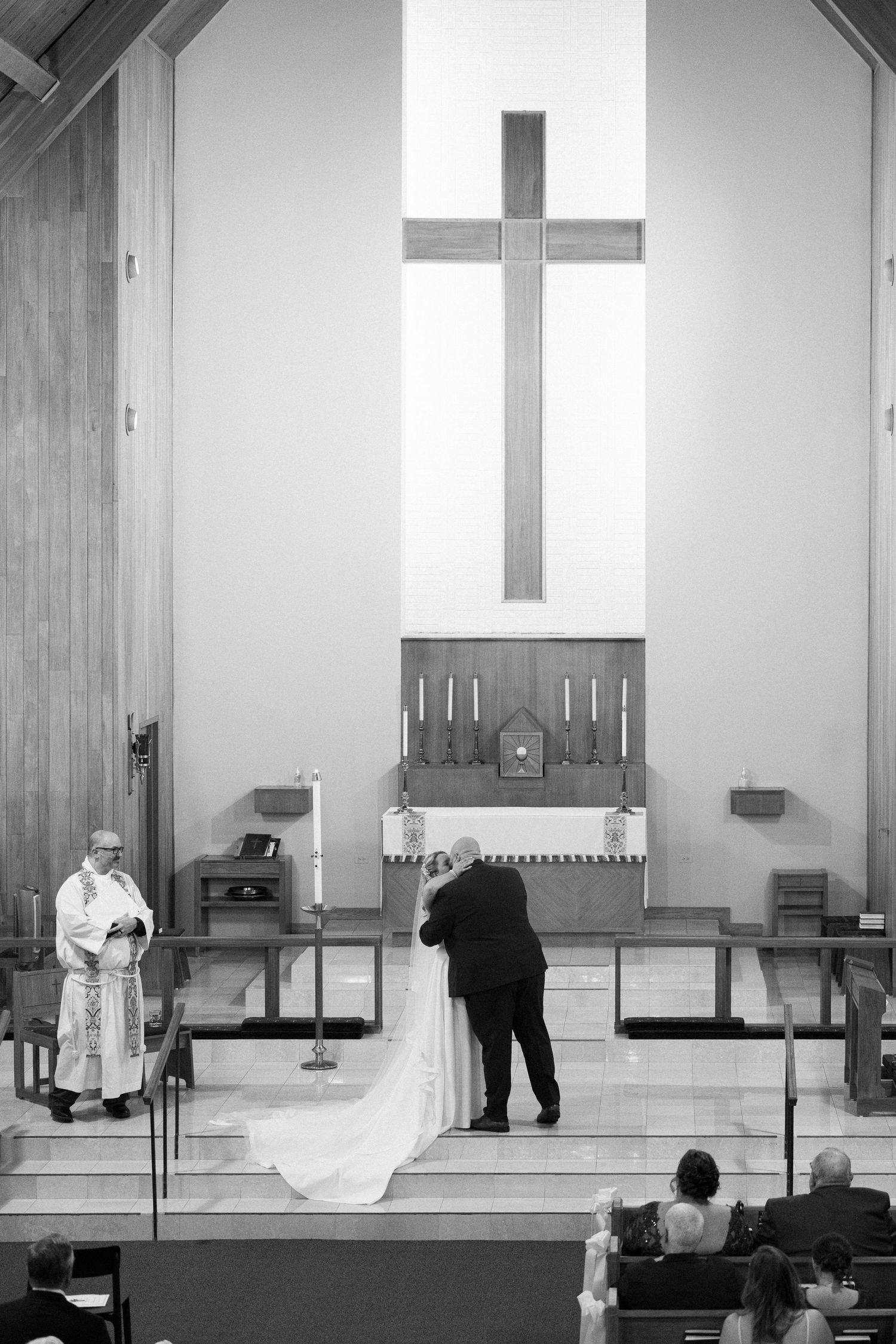 Black and white photo of a couple sharing their first kiss at a church wedding altar with a large cross behind them.