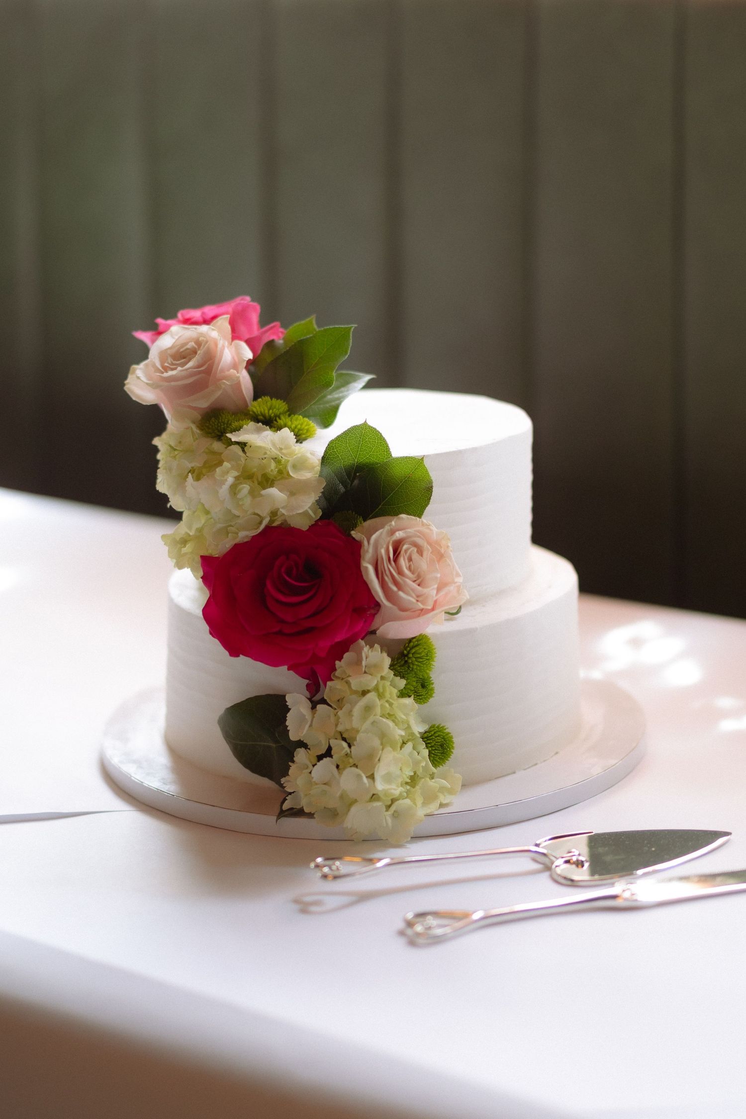 Two-tier white wedding cake adorned with pink and red roses and green leaves, displayed with silver cake serving utensils.
