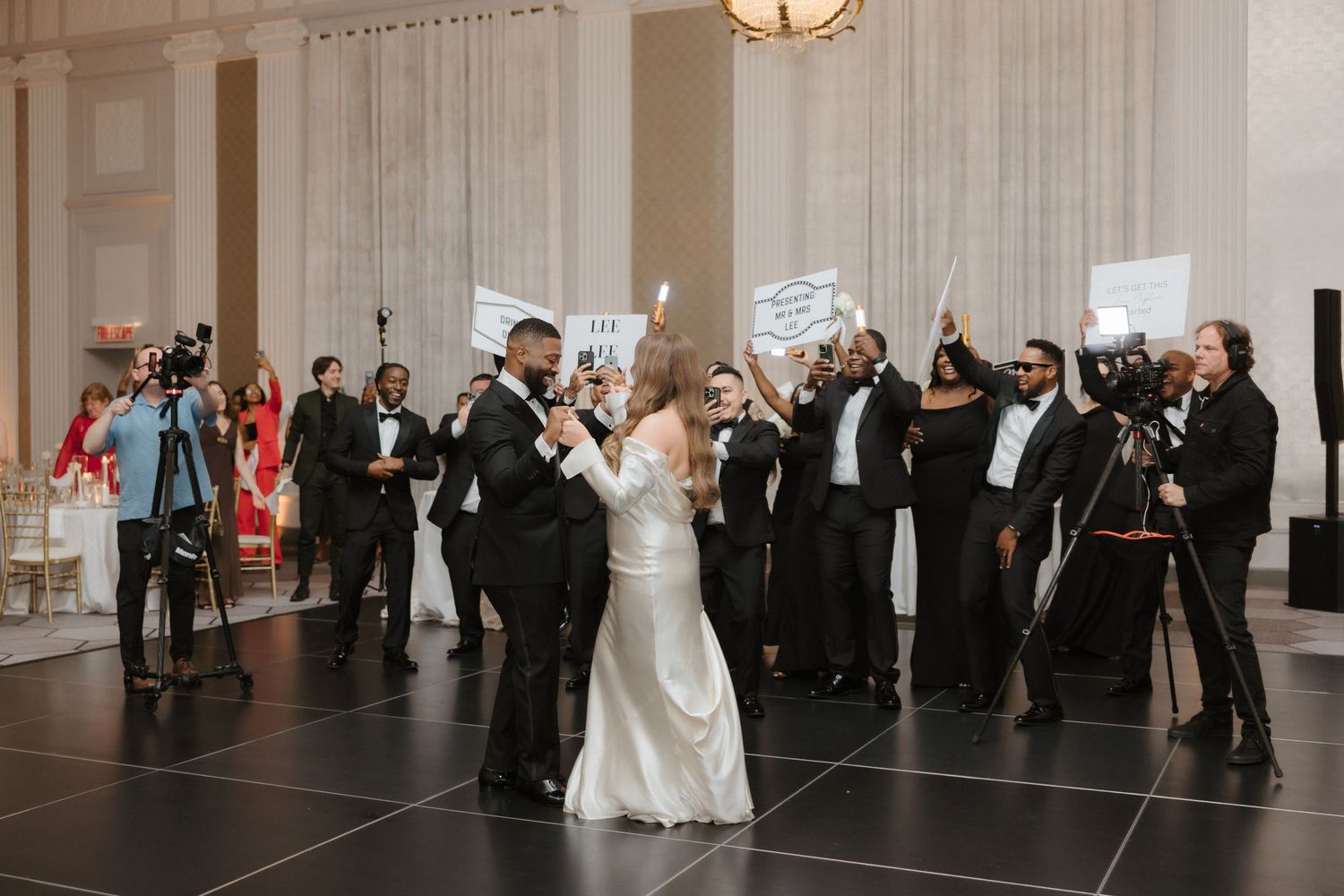 Bride and groom share first dance surrounded by guests holding signs at elegant wedding reception.