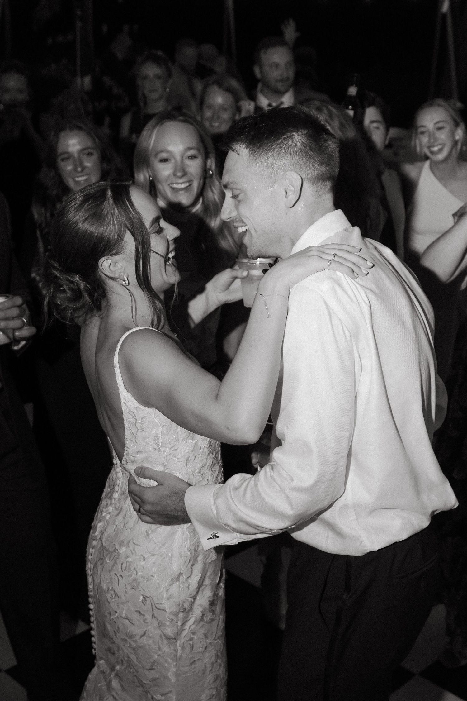 Black and white photo of bride and groom sharing a joyful first dance surrounded by smiling wedding guests.
