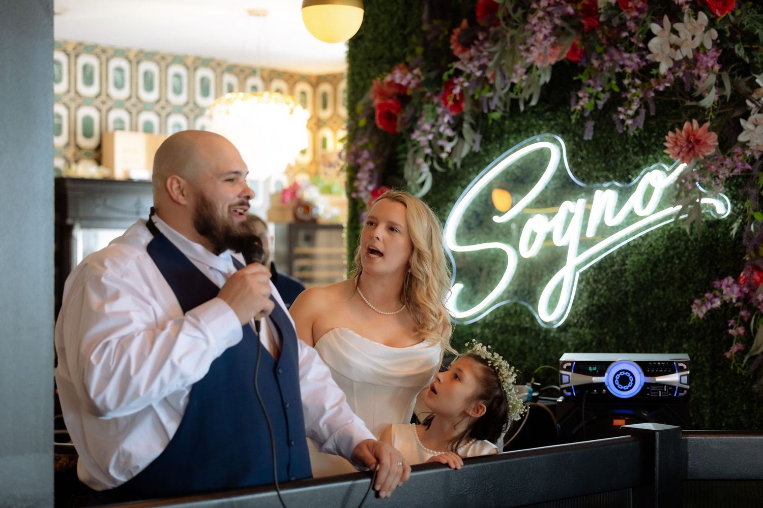 Groom speaks into microphone beside bride under glowing Sogno sign at elegant wedding reception venue.