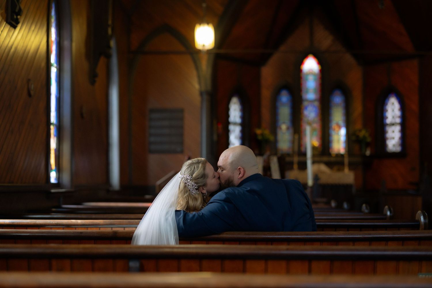Bride and groom sharing a tender kiss while seated in wooden church pews, with stained glass windows in the background.