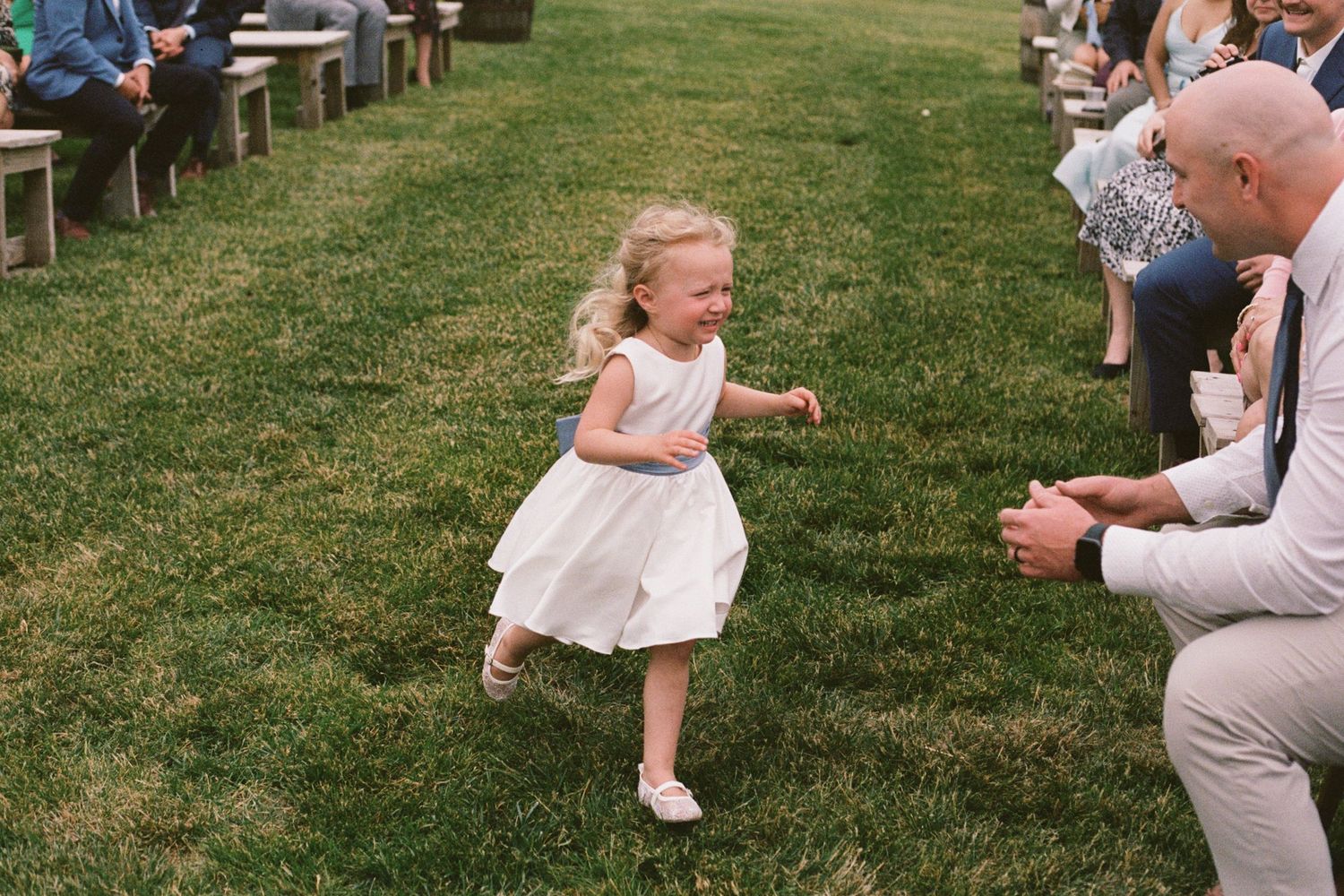 A joyful flower girl in a white dress runs down the grassy aisle at an outdoor wedding ceremony.