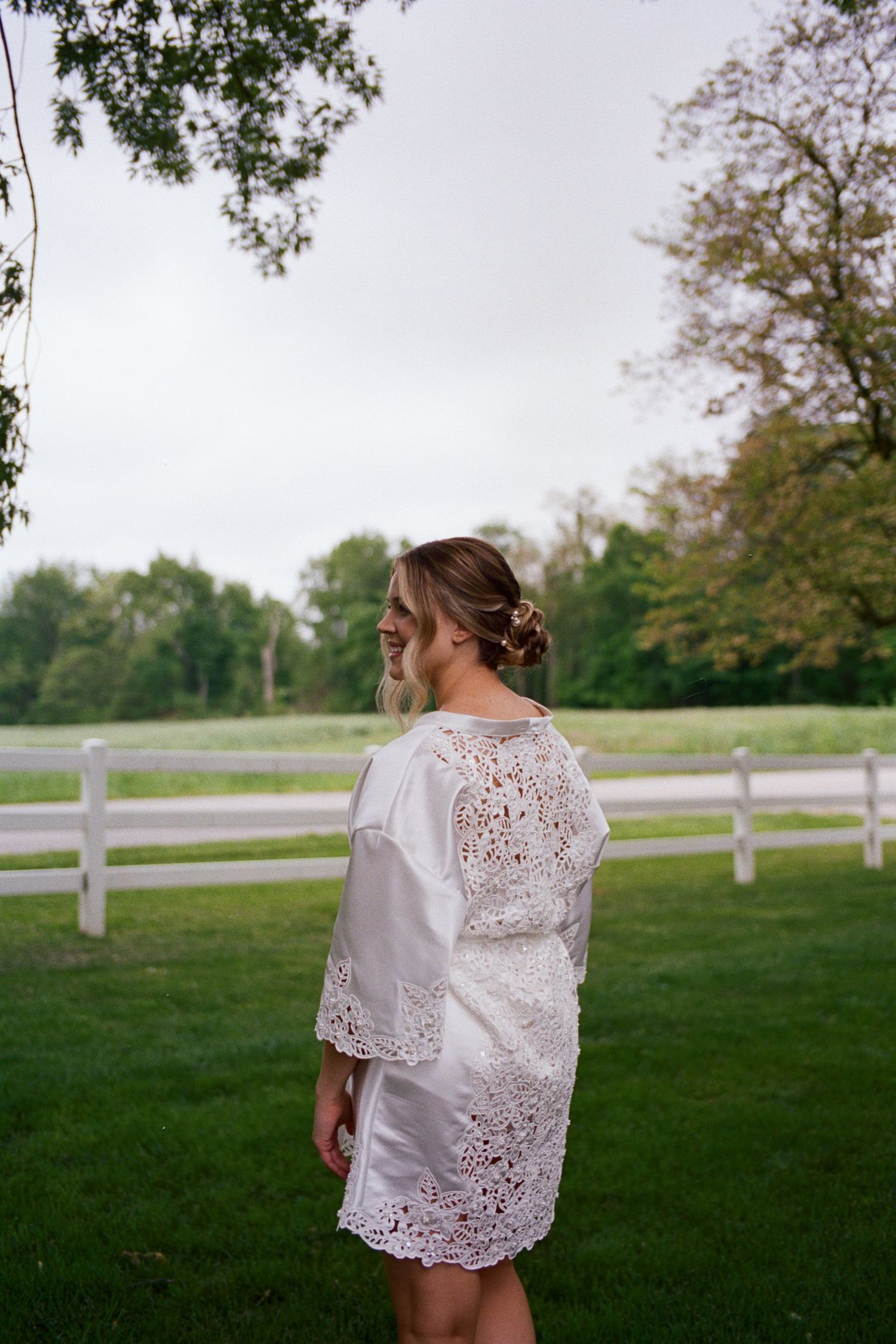 Woman in white lace dress standing on green lawn near white fence, smiling, with trees in background.